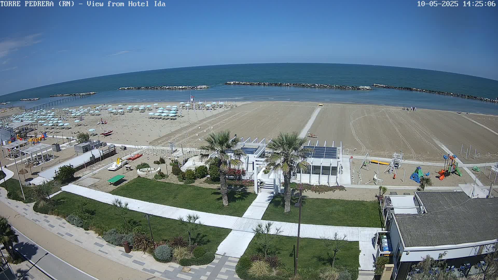 An aerial view of a beach on a sunny day shows a sandy shore with light blue umbrellas, a playground, and a walkway leading to a beachfront building.