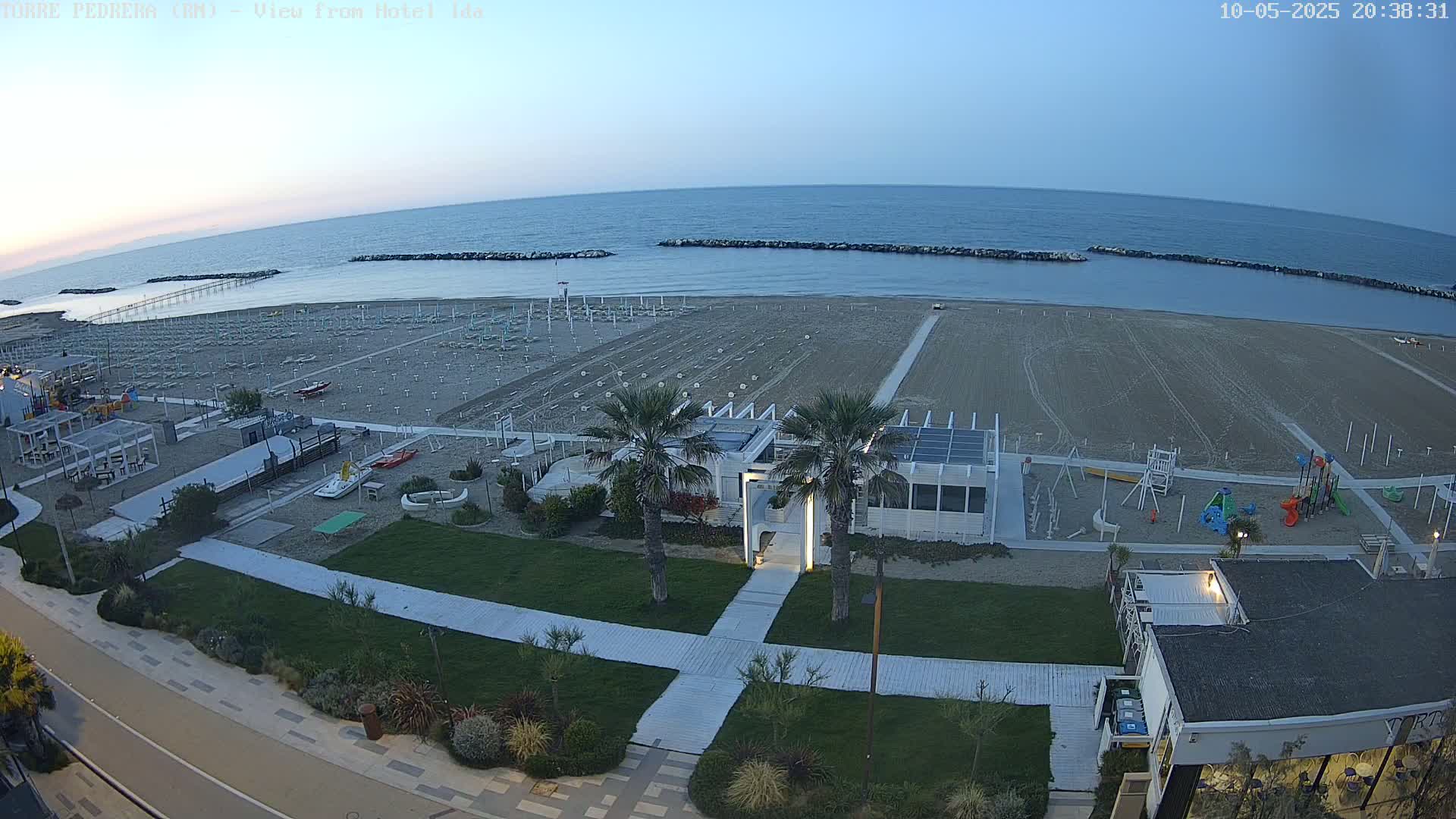 An empty sandy beach at dusk, with rows of beach umbrellas folded up, overlooks a calm sea, and features a playground and beachfront buildings.
