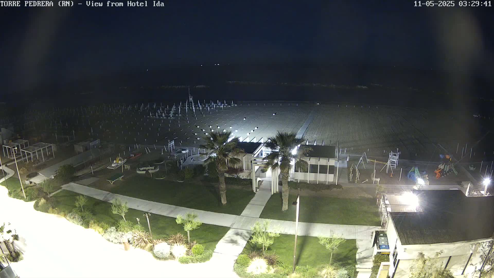 A nighttime, overhead view shows a mostly empty sandy beach with closed umbrellas and a playground, adjacent to a grassy area with palm trees and buildings under artificial light.
