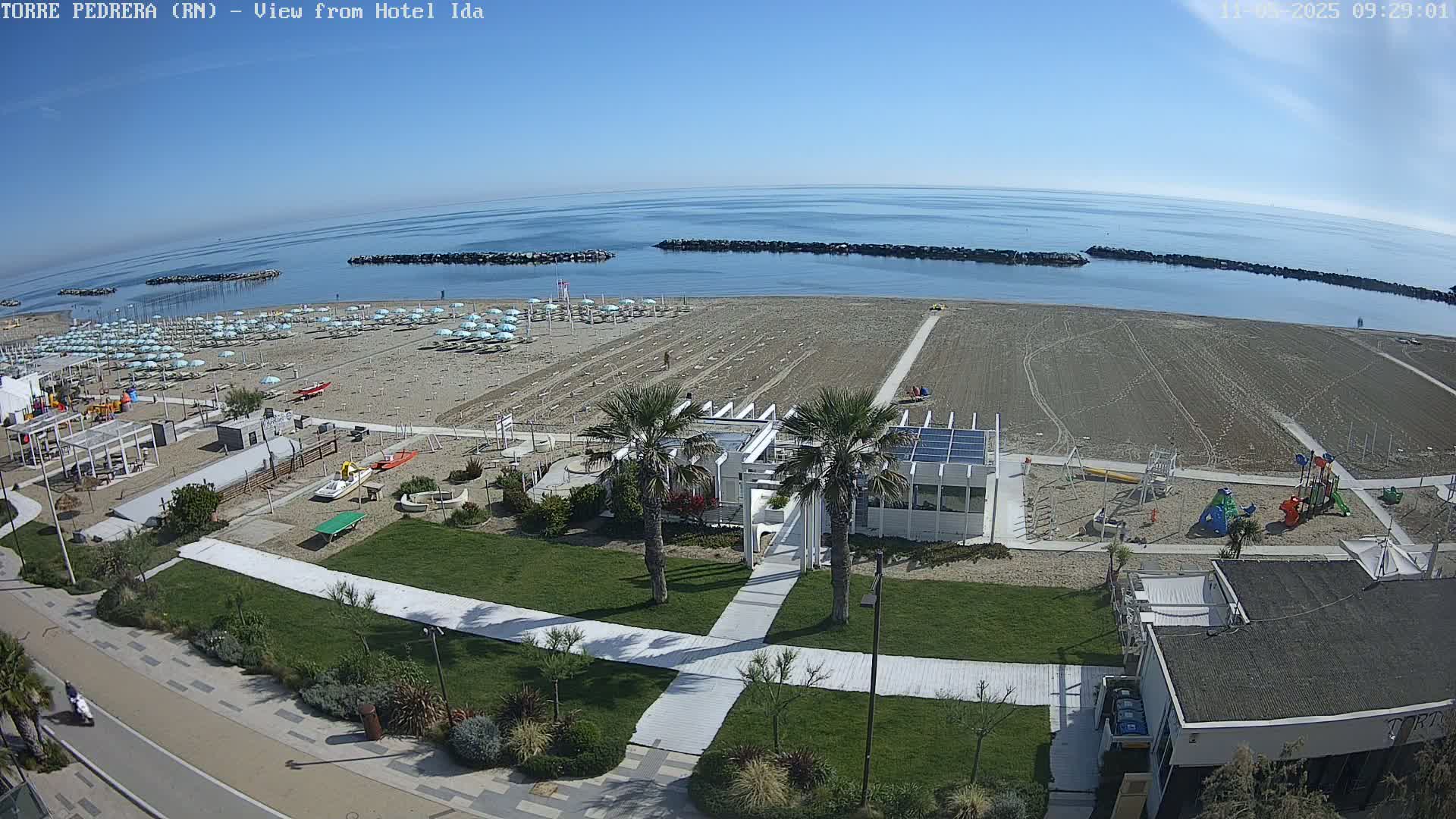 A sandy beach with rows of beach umbrellas, a playground, and palm trees is shown on a clear, sunny day, with calm ocean water visible in the background.