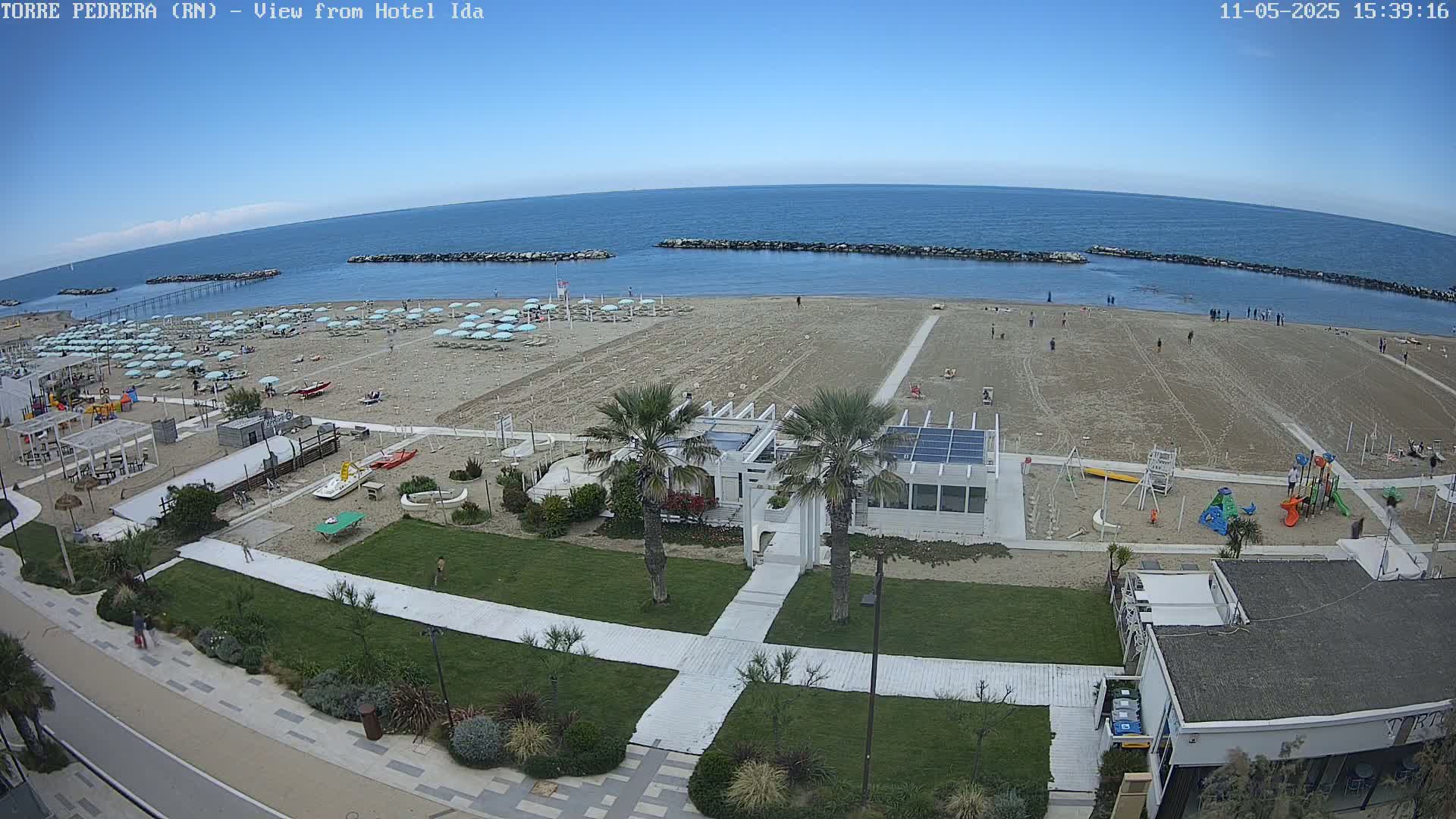 An aerial view shows a beach with light-blue umbrellas, a playground, and a white building under a clear blue sky.