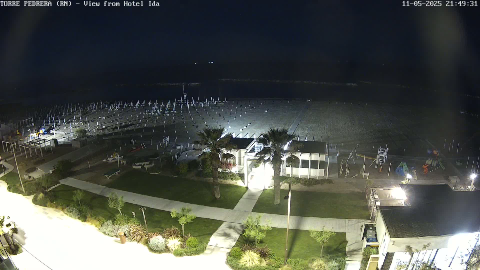 A nighttime aerial view shows an empty beach with closed umbrellas, a playground, and a building with palm trees, all illuminated under a dark sky.