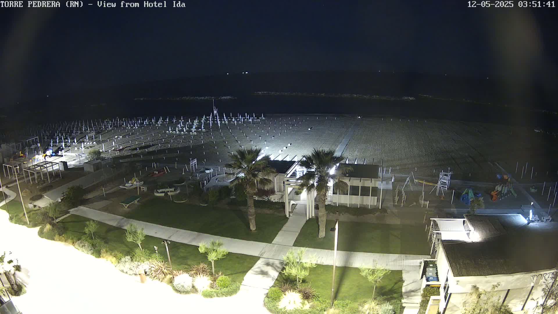 A nighttime view of a beach with rows of closed umbrellas, a small building, a playground, and palm trees, under a clear, dark sky.