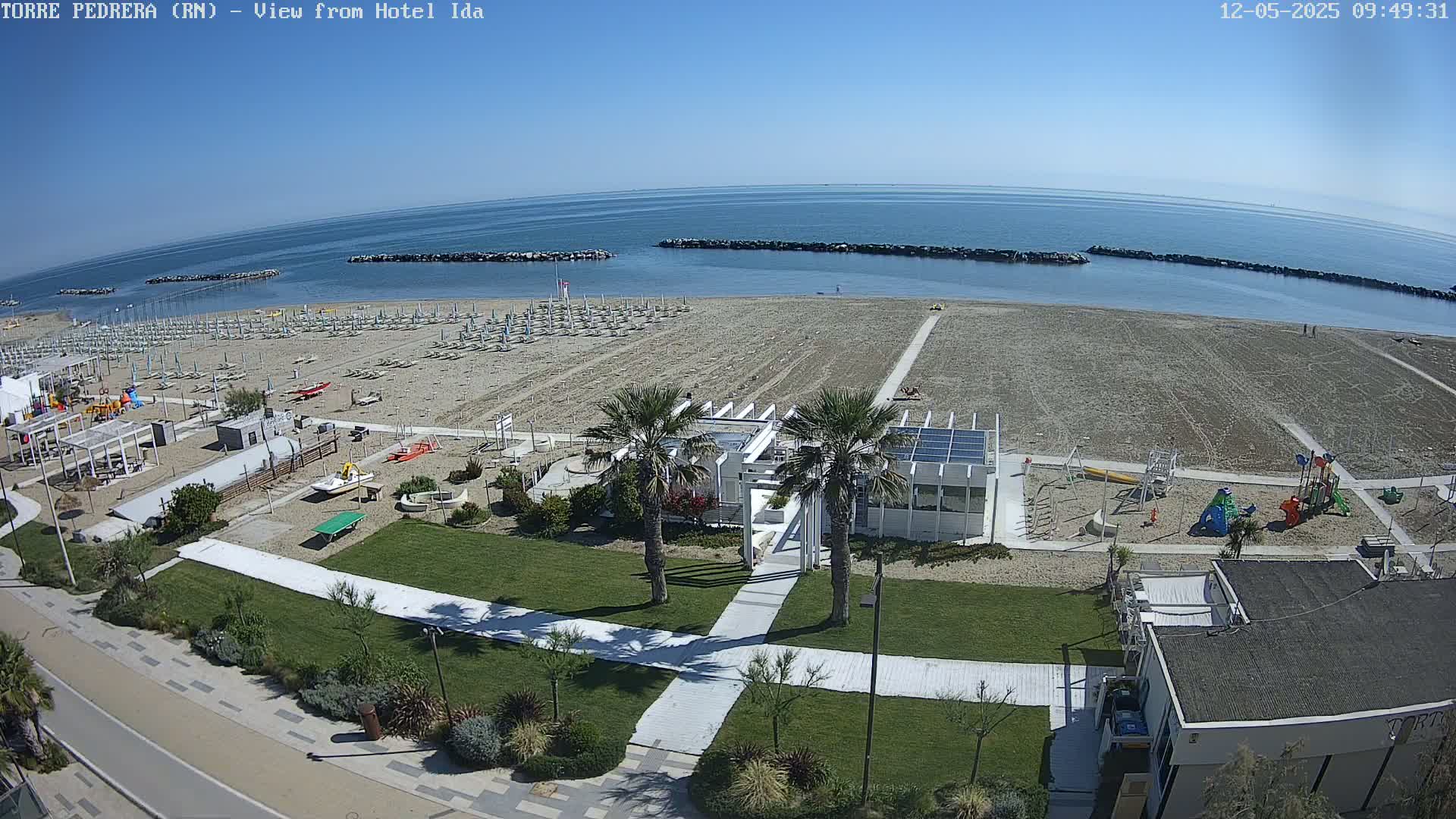 A sandy beach with rows of beach umbrellas, a playground, and palm trees is visible on a sunny day, with a calm ocean in the background.