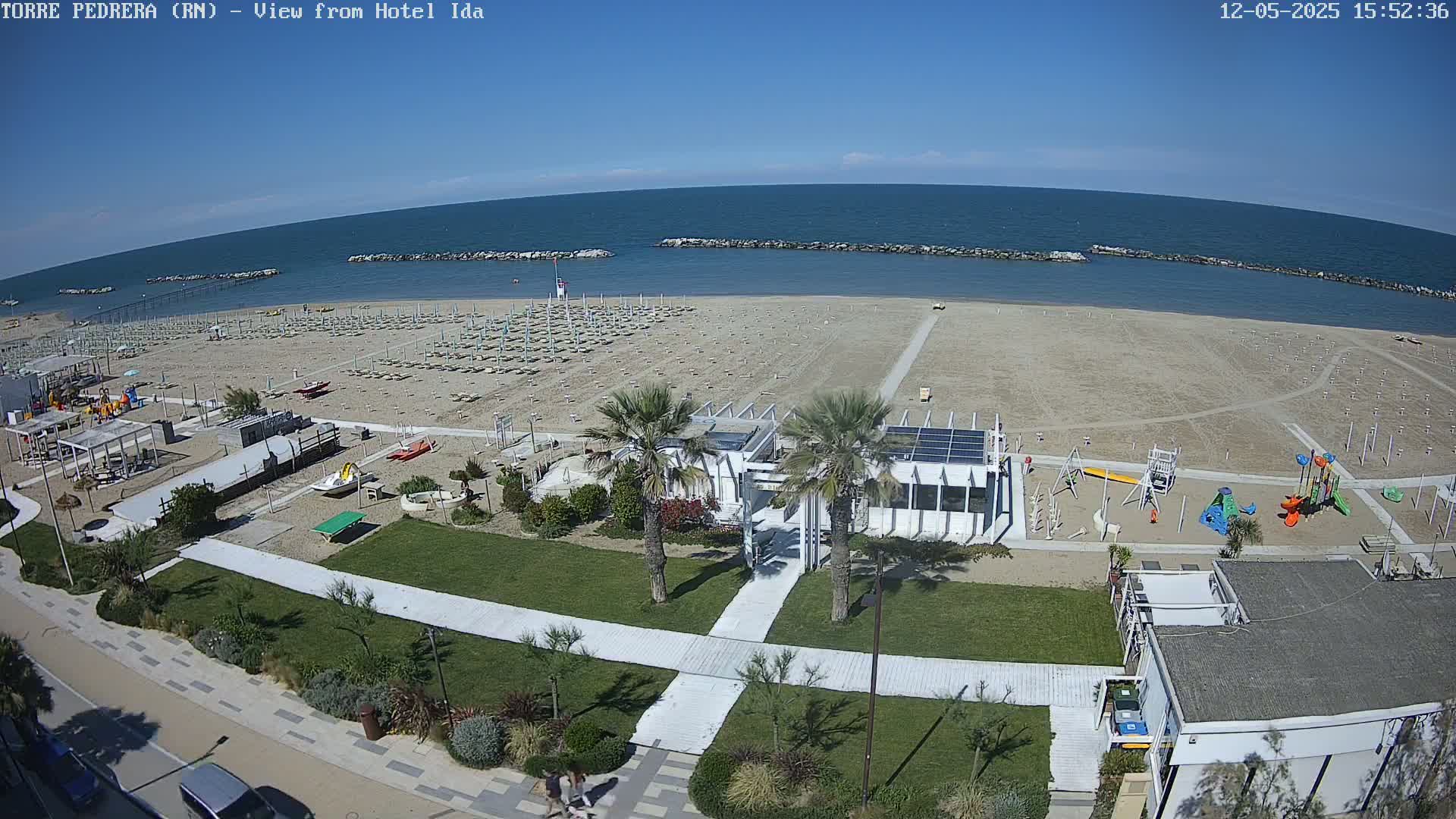 A beach with rows of beach umbrellas and lounge chairs, children's playground equipment, and a beachfront building, all under a clear blue sky.