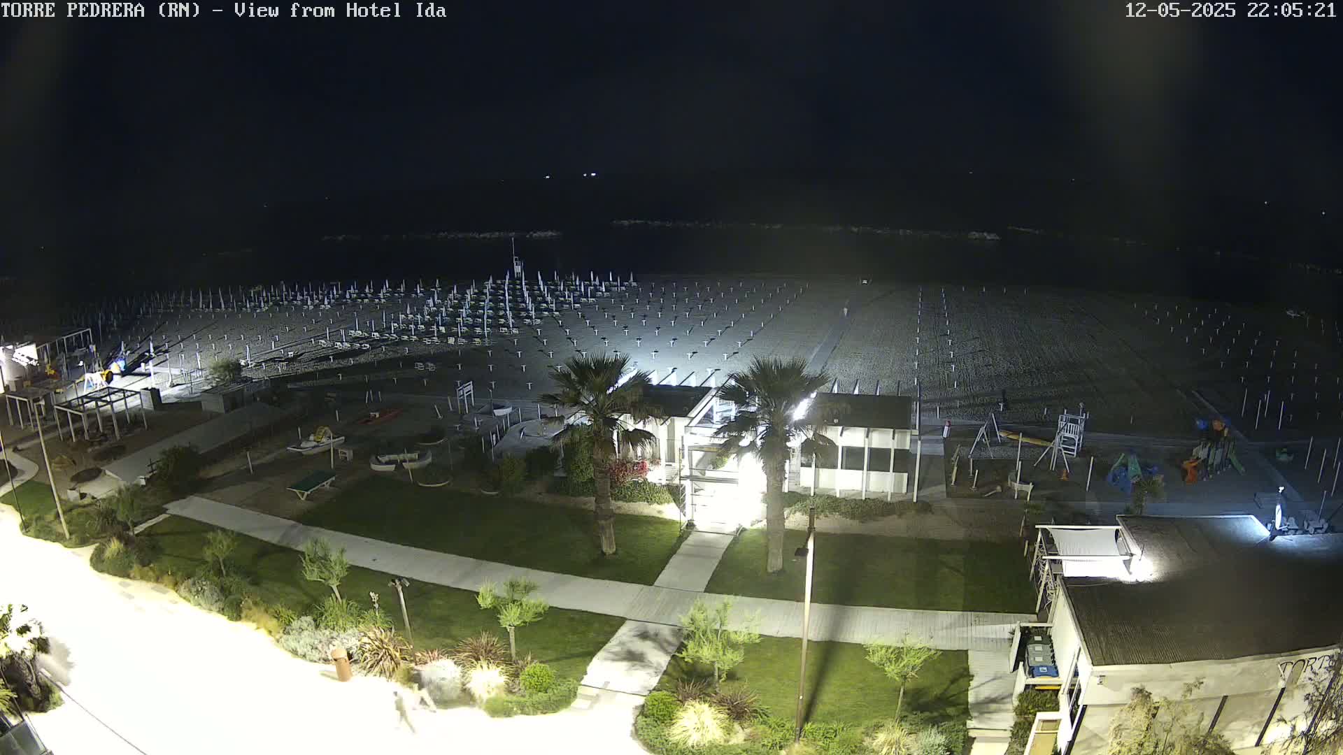A nighttime view of a beach with rows of closed beach umbrellas, a playground, and low-light landscaping surrounding a building.
