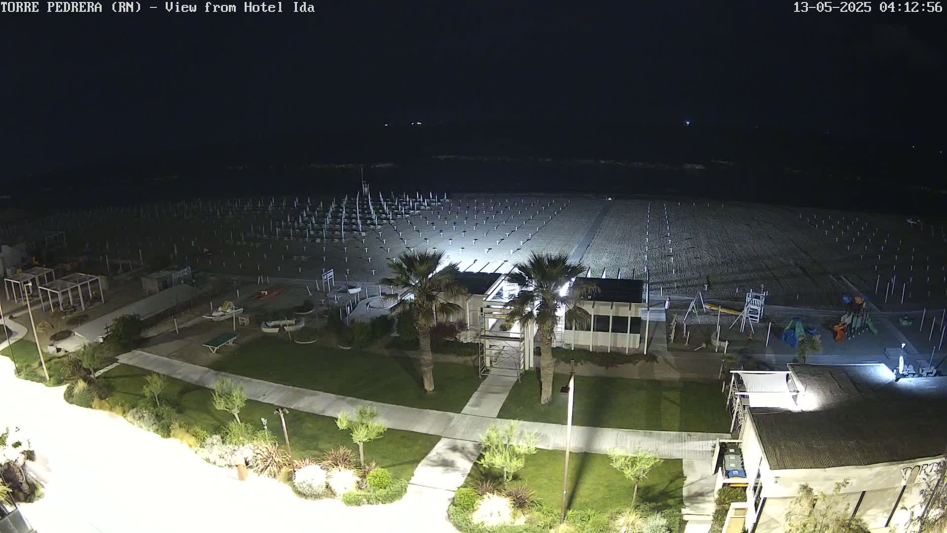 A nighttime view of a mostly empty beach with rows of beach umbrellas and a playground, adjacent to a building and landscaped area.