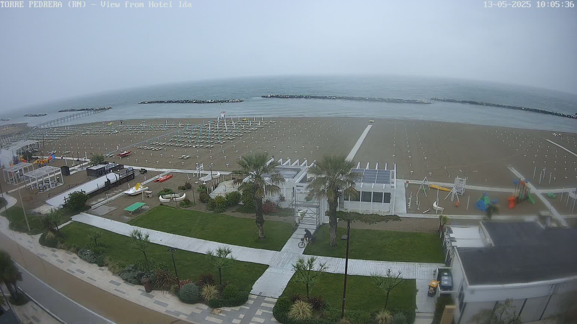A mostly empty beach with rows of unoccupied beach chairs and umbrellas under an overcast sky is seen from above, along with a nearby playground and boardwalk.
