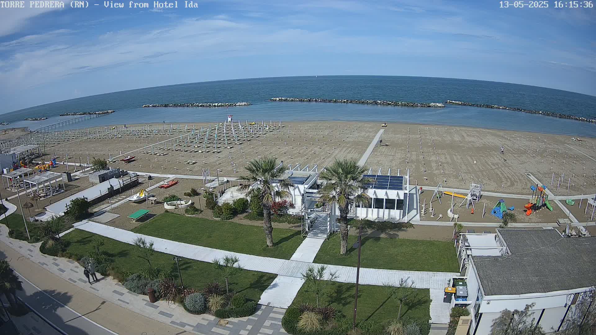 A sandy beach with rows of beach umbrellas, a playground, and a beachfront building is visible under a mostly sunny sky, with calm ocean water beyond.