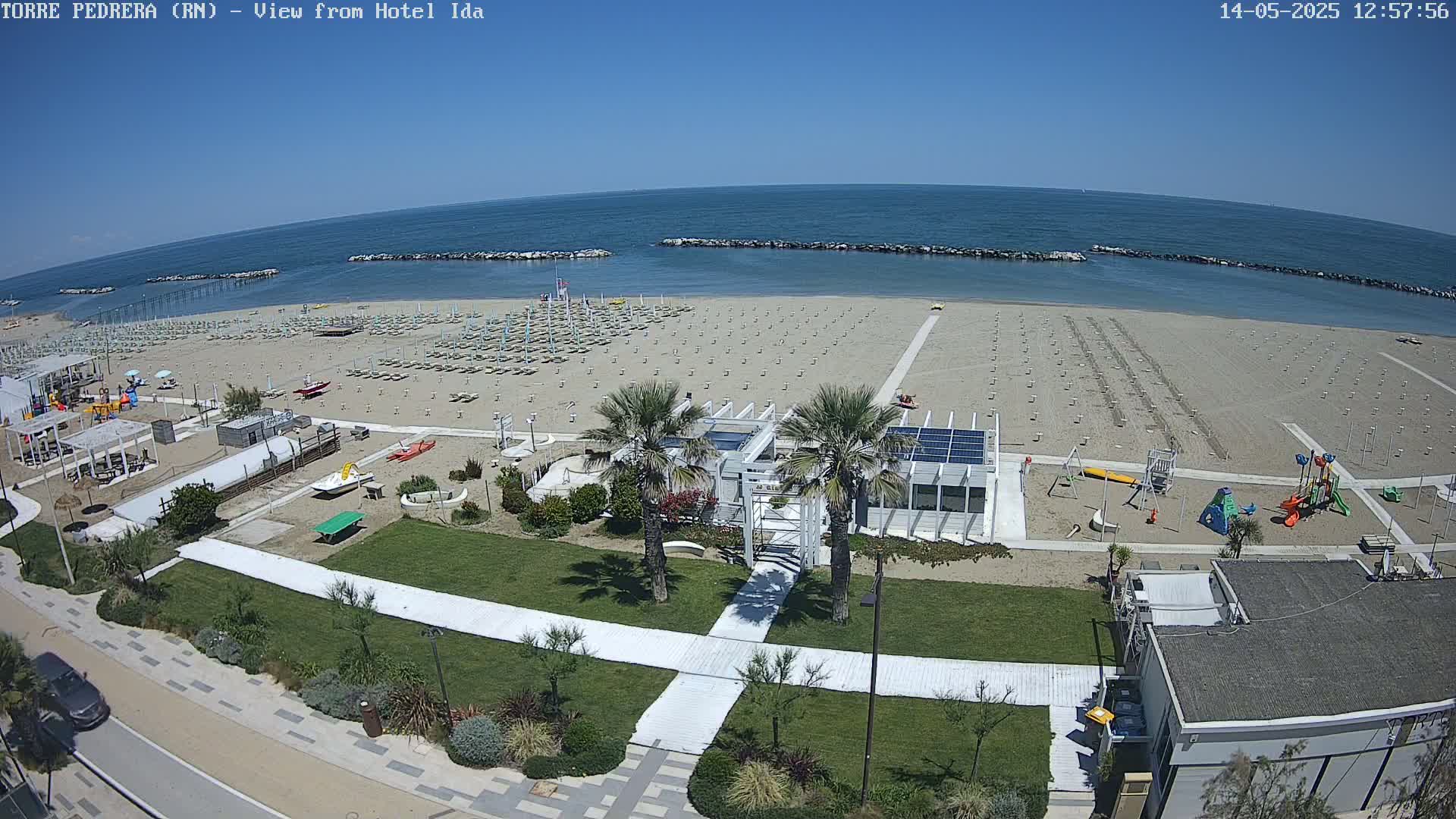 A sunny beach with rows of beach umbrellas, a playground, and palm trees near a building, all overlooking a calm ocean.