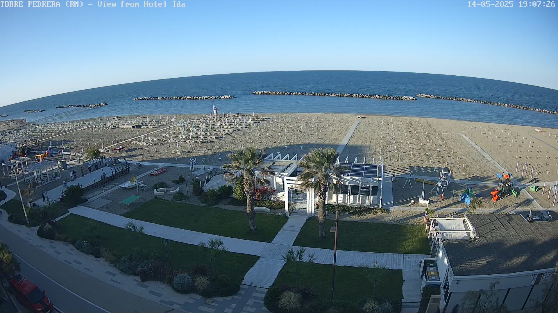 A sandy beach with rows of closed umbrellas, a playground, and a beachfront building is seen on a clear, sunny day, with calm blue water stretching to the horizon.
