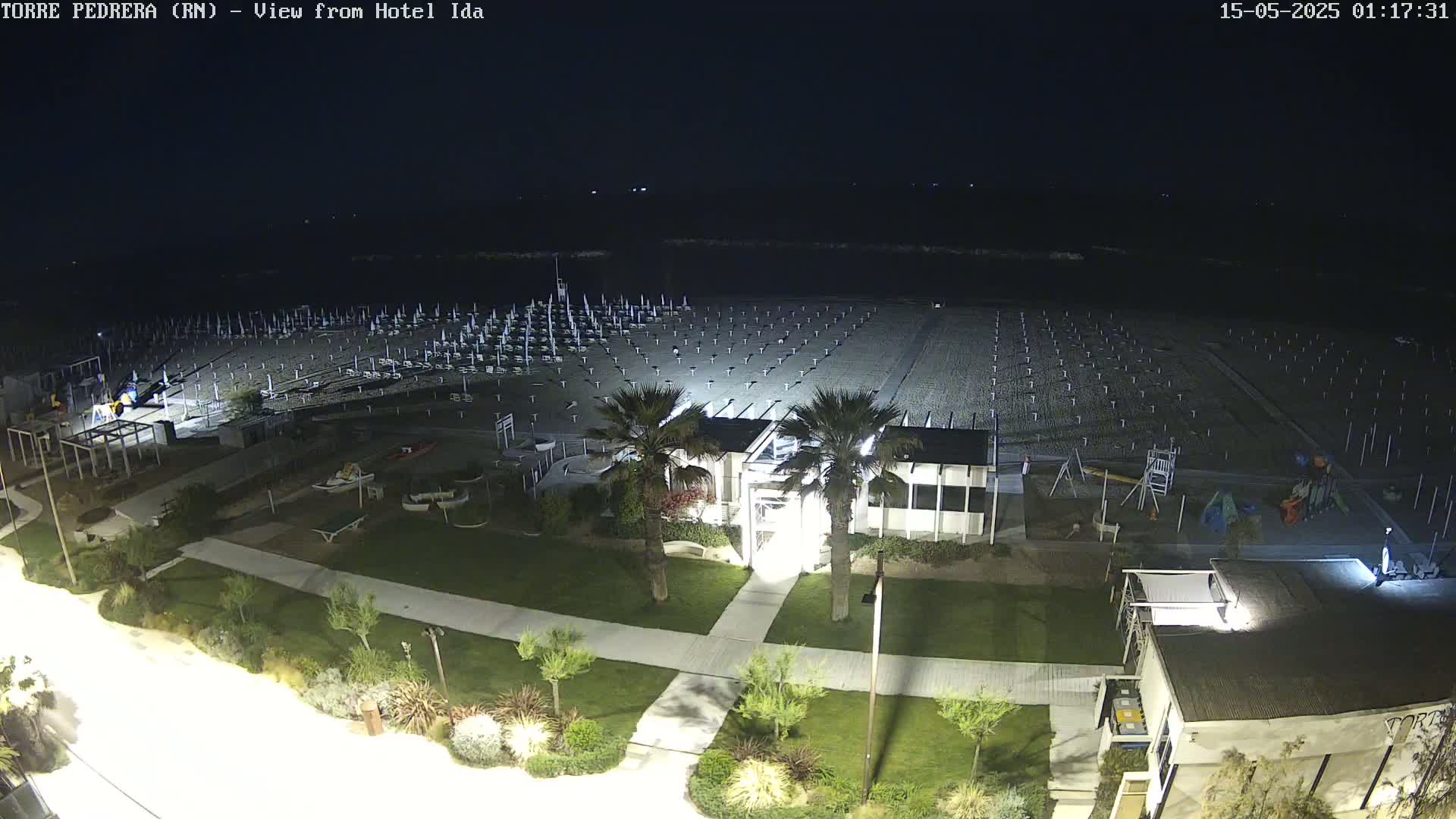 A nighttime, overhead view shows a mostly empty beach with rows of closed umbrellas, a playground, and beachfront buildings illuminated under dark skies.