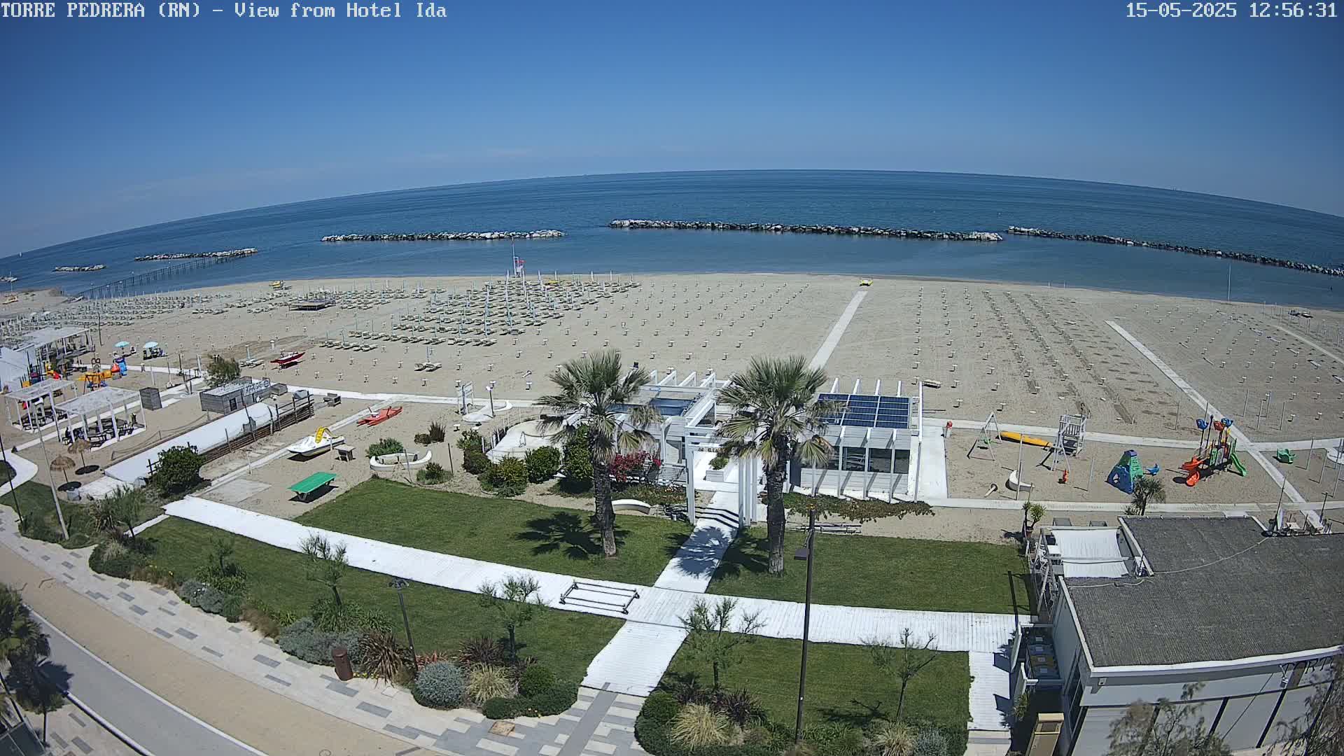 An aerial view of a beach with rows of empty beach chairs under a clear, sunny sky, showing a playground and beachside building.