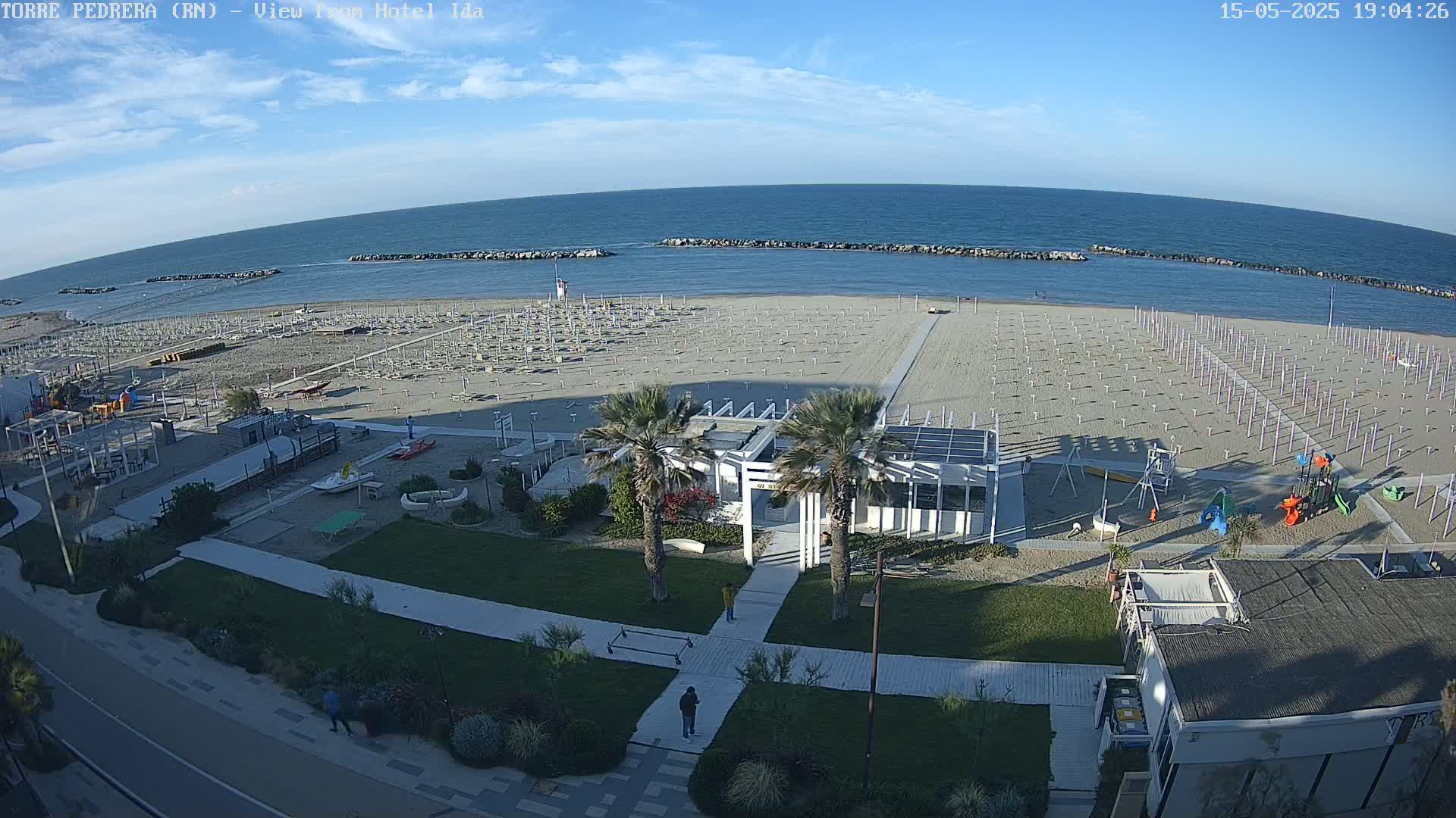 An aerial view shows a beach with numerous empty umbrellas and chairs, a playground, and a beachfront building, all under a mostly sunny sky.