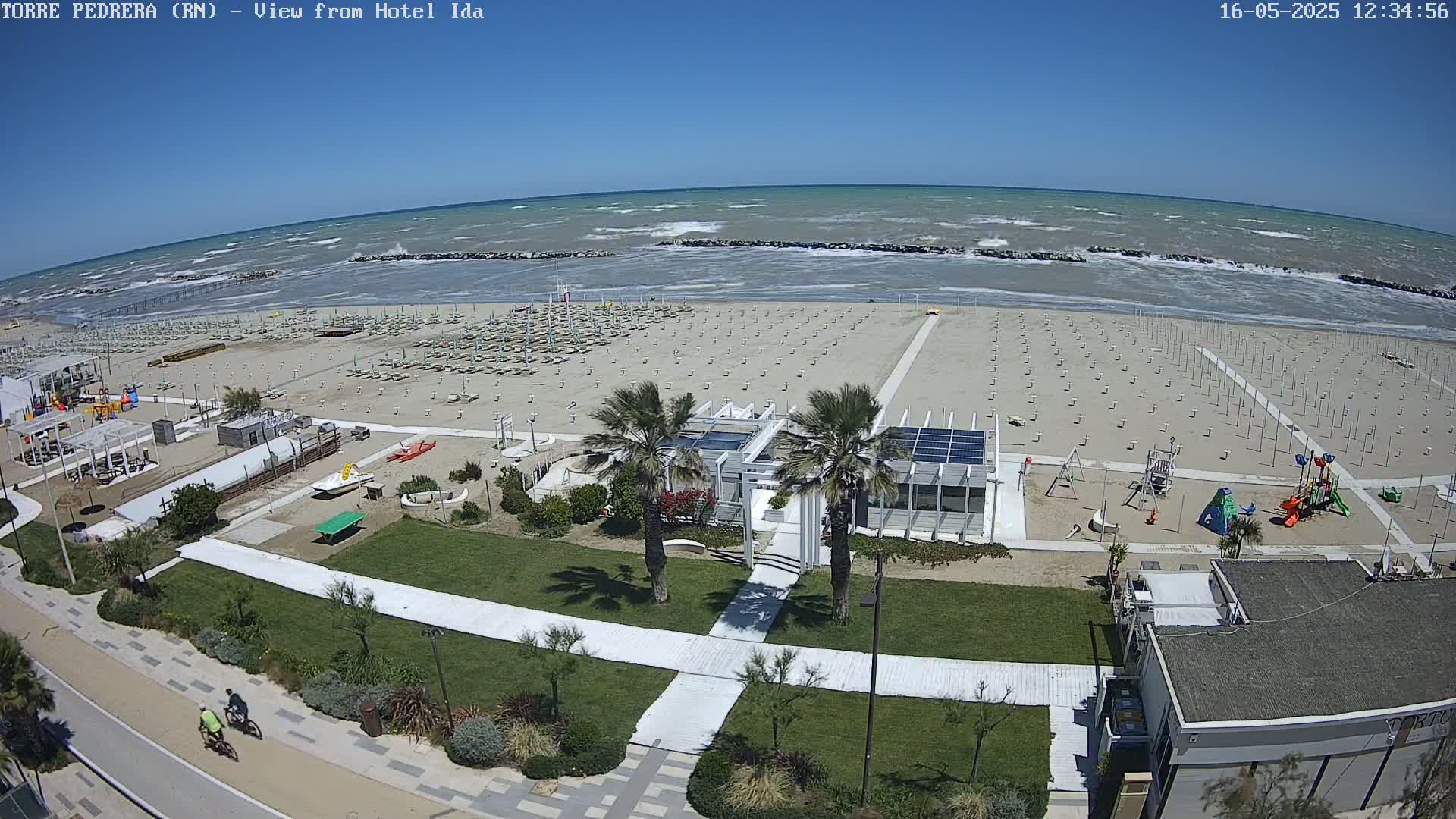 A mostly sunny day at a beach shows rows of empty beach umbrellas on the sand, a children's playground, a beachfront building, and two cyclists on a paved path next to a windy sea with moderate waves.