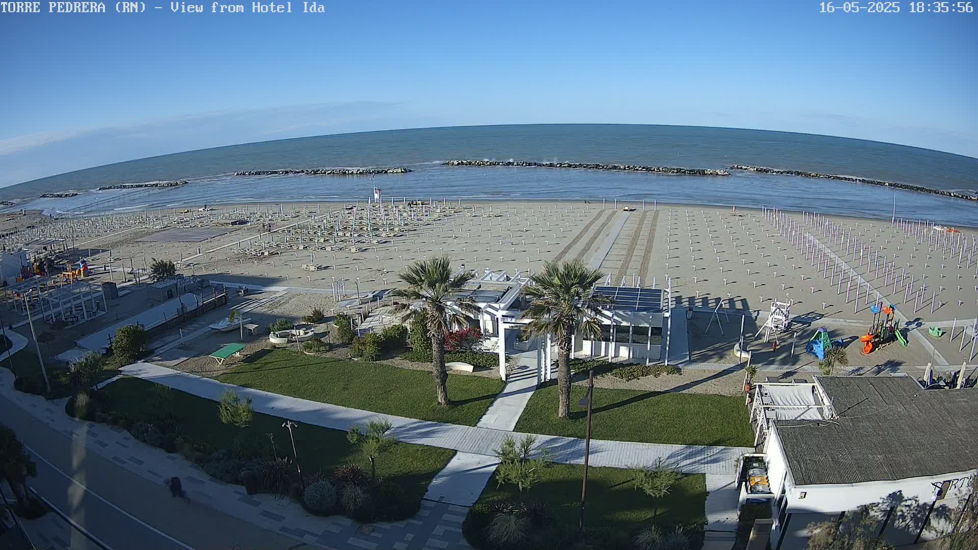 A beach with numerous closed beach umbrellas, a playground, and a small building is visible under a clear blue sky.