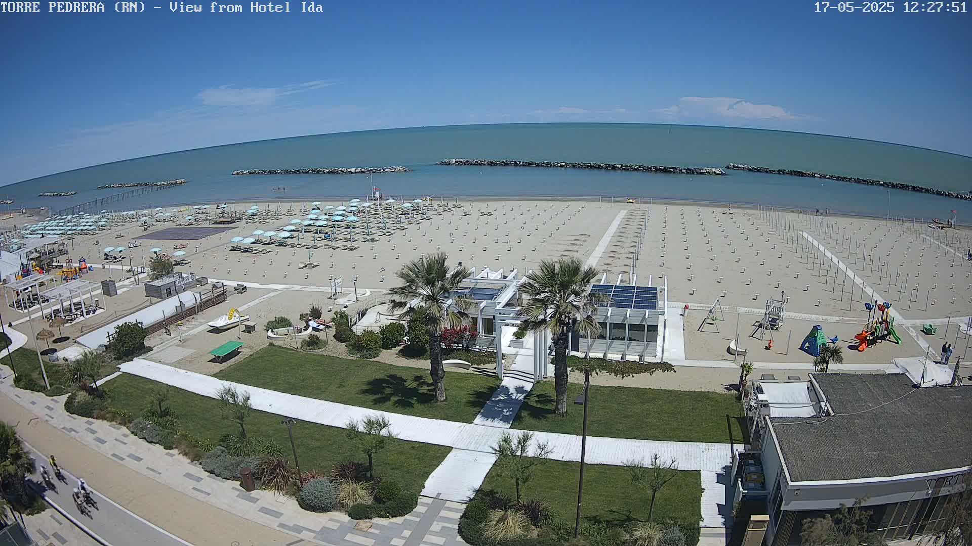 An aerial view shows a beach with many empty umbrellas and a playground under a clear blue sky.