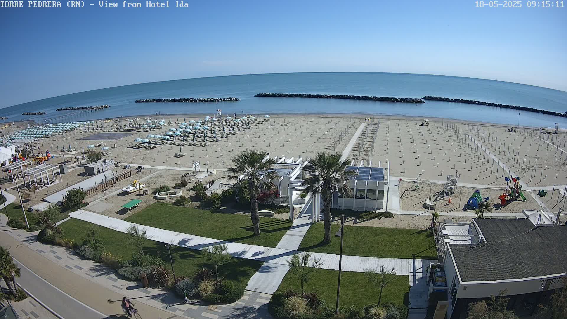 A sandy beach with rows of closed umbrellas, a beachfront building, and a playground is seen from above on a sunny day, next to a calm ocean.