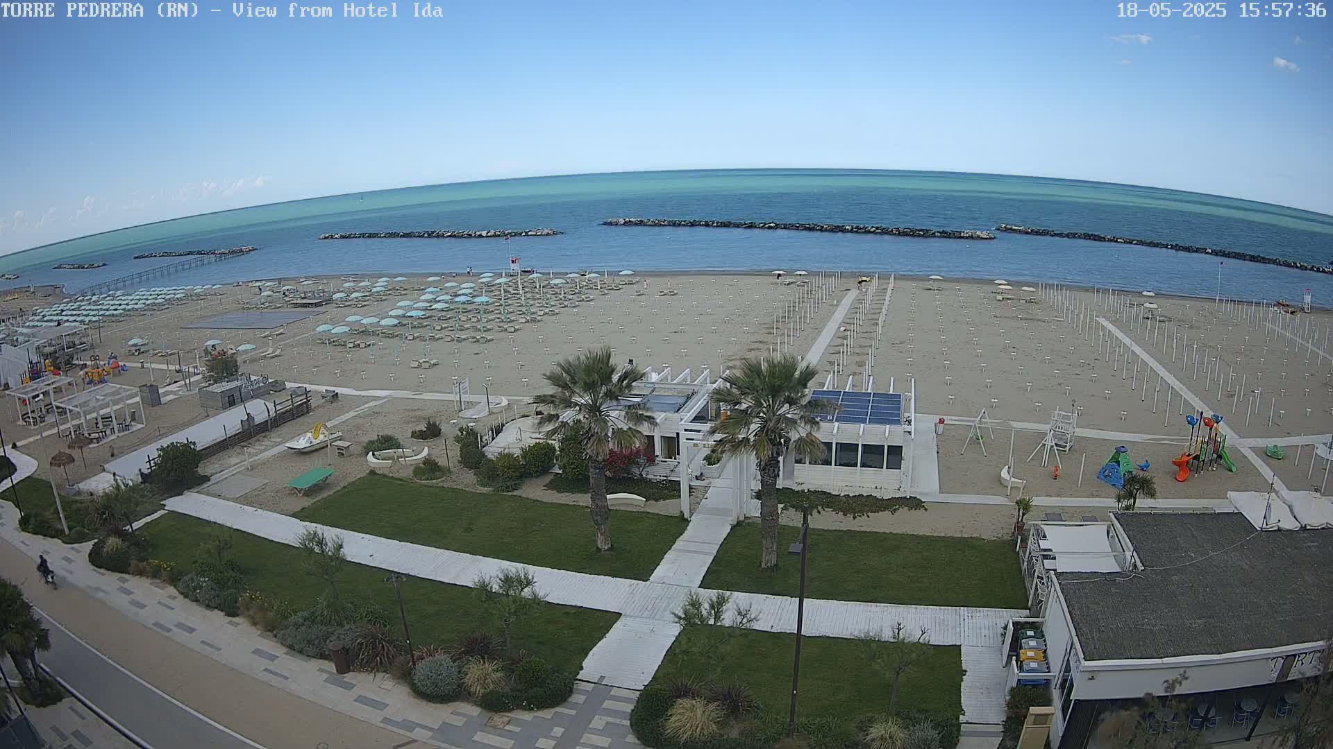 An aerial view shows a beach with numerous umbrellas and beach chairs under a clear blue sky, adjacent to a grassy area with palm trees, walkways, and a playground.