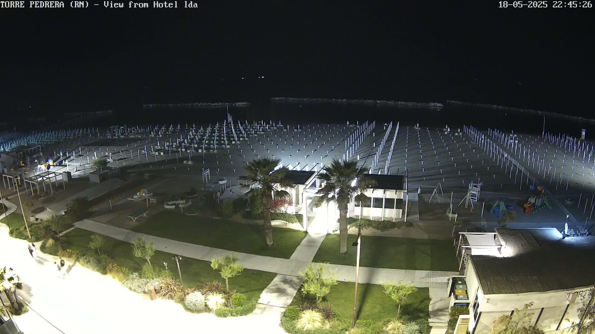 A nighttime aerial view of an empty beach with rows of closed umbrellas and beach chairs, adjacent to a grassy area with landscaping and a building.