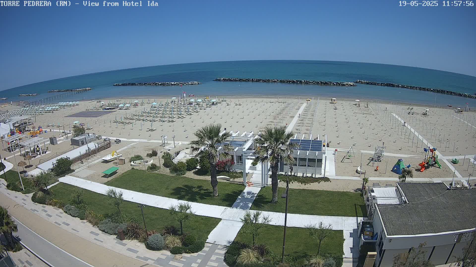 An aerial view of a beach on a sunny day shows rows of beach umbrellas and lounge chairs, a playground, and a beachfront building with palm trees.