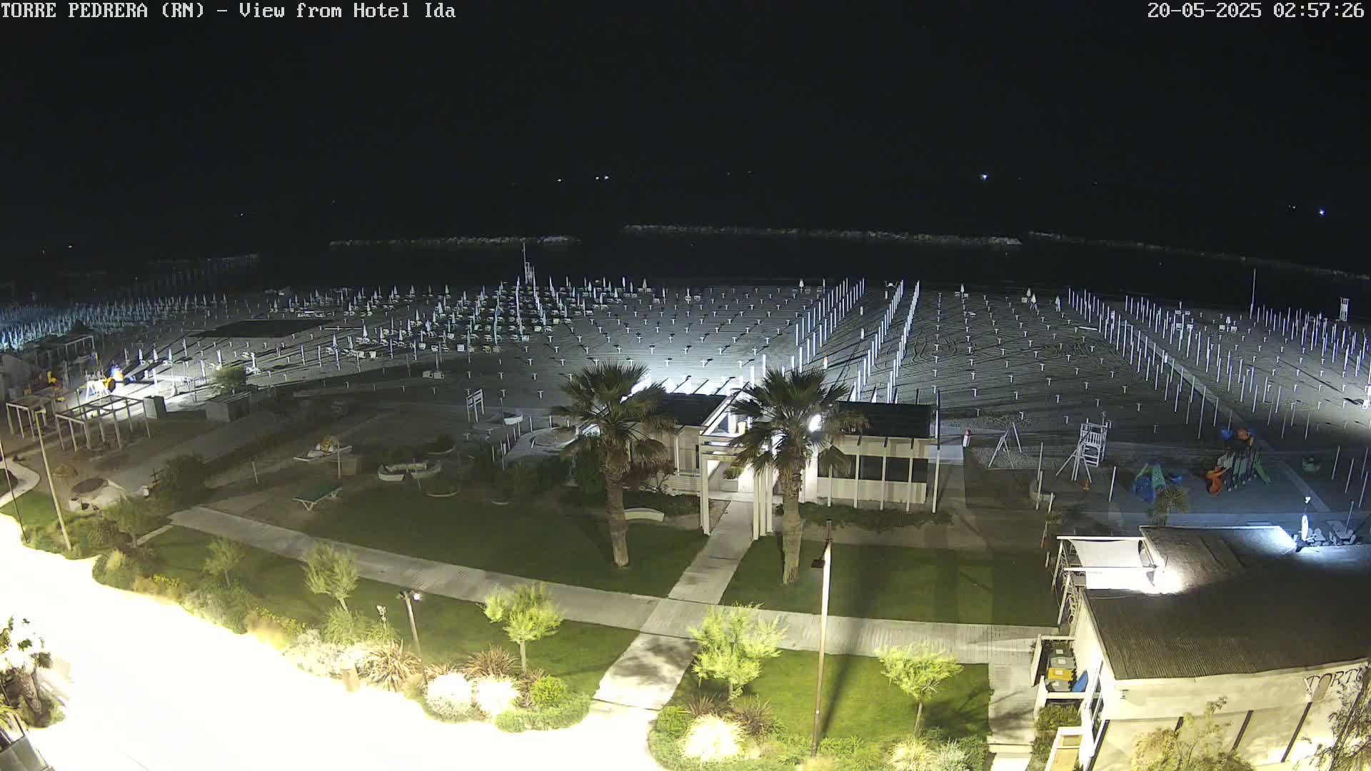 A nighttime, overhead view of a beach with rows of beach umbrellas and chairs, a small building, playground equipment, and landscaping.