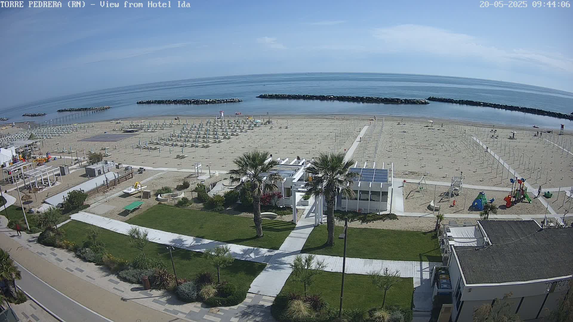 An overhead view shows a mostly empty sandy beach with rows of unoccupied beach umbrellas, a playground, and a small building under a clear, sunny sky.