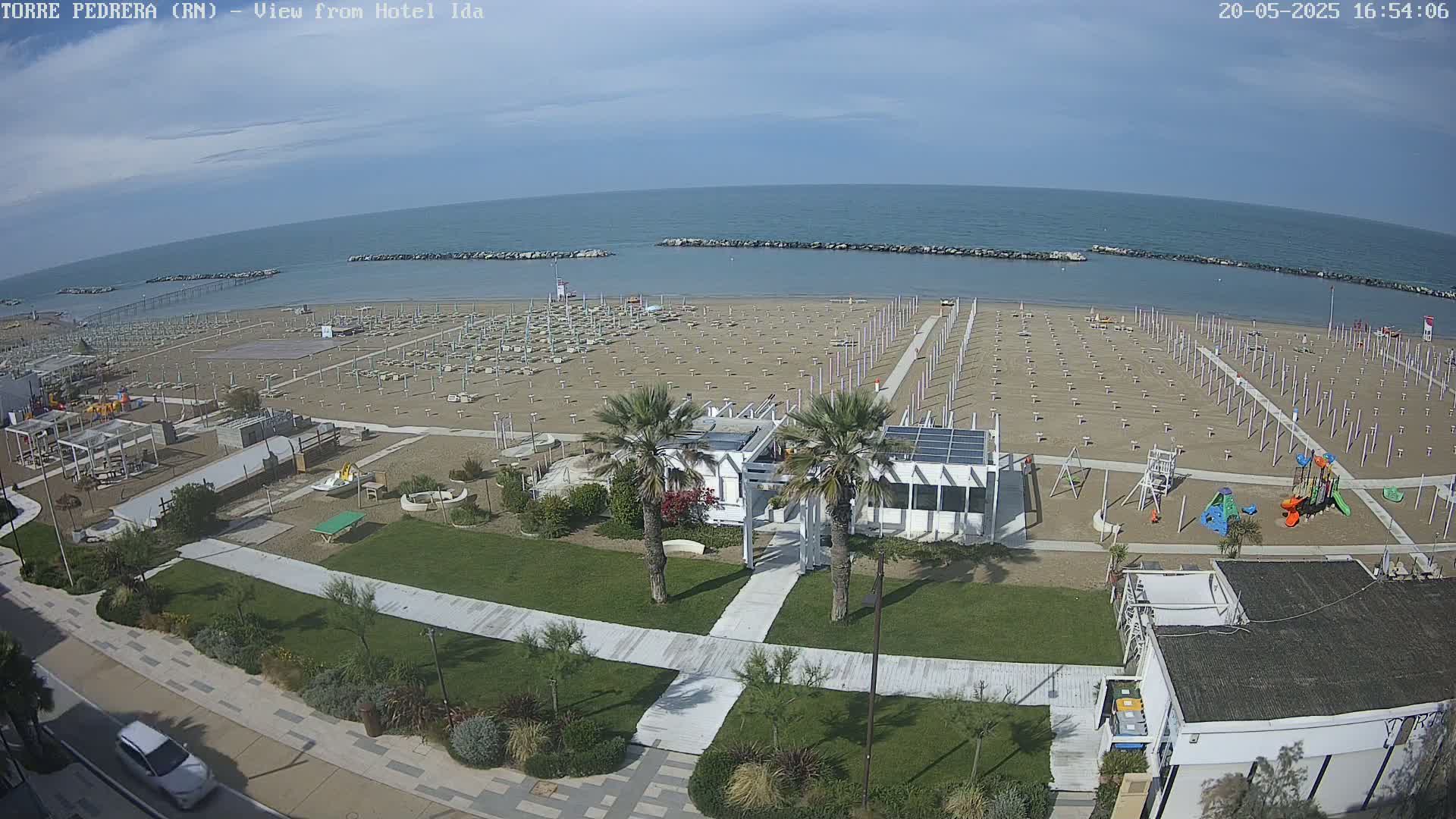 An aerial view shows a sandy beach with rows of umbrellas and beach chairs, a playground, a small building, and palm trees, all under a mostly clear sky.