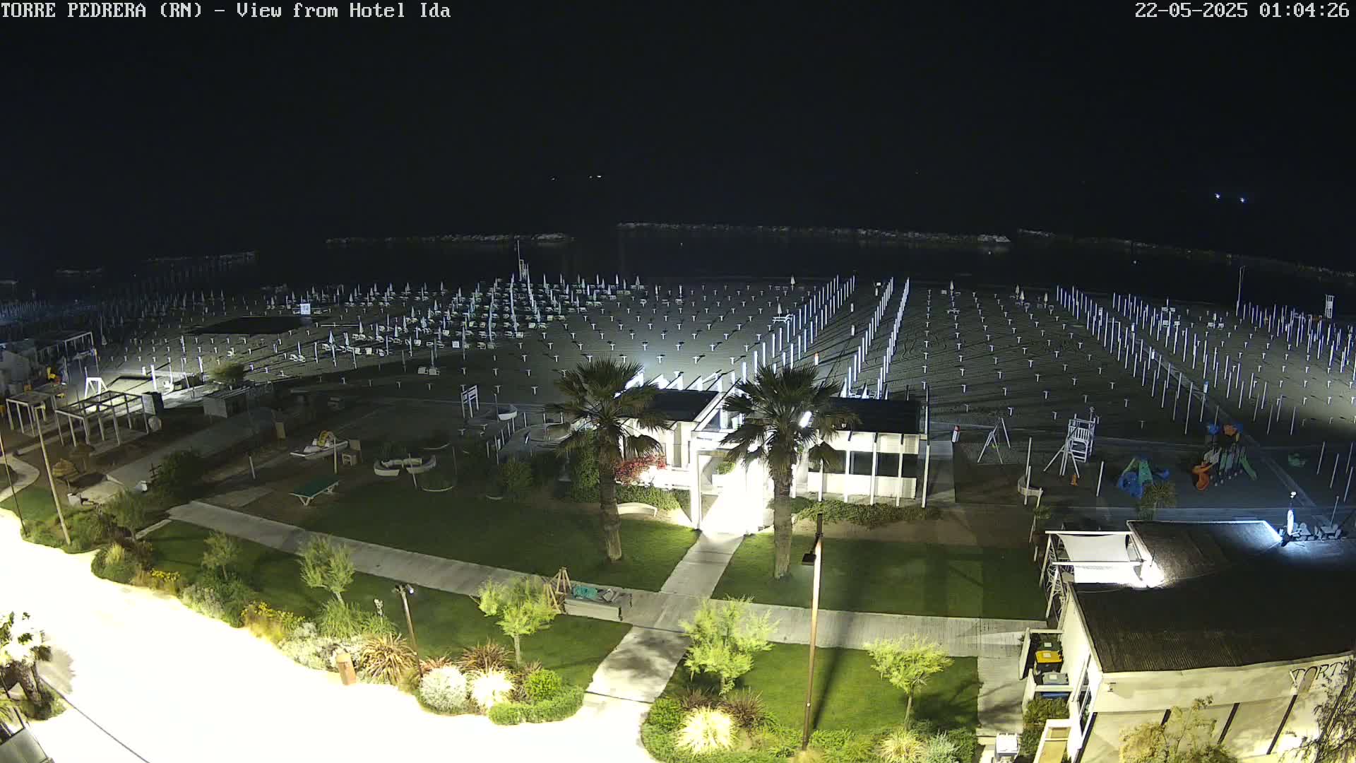 A nighttime aerial view of a beach with rows of empty beach umbrellas and chairs, a playground, and some nearby buildings, under a clear dark sky.