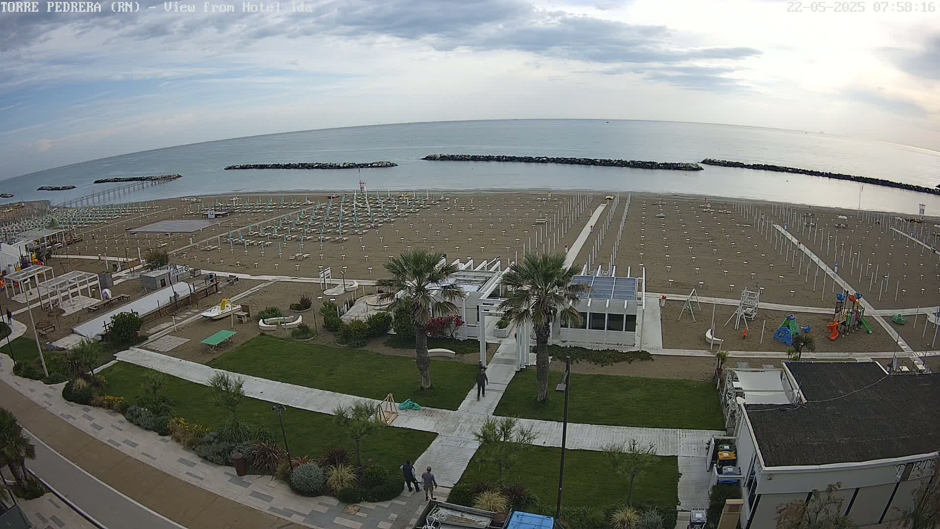 An overhead view of a sandy beach with rows of unoccupied beach umbrellas and chairs, a playground, and a calm ocean under a partly cloudy sky.