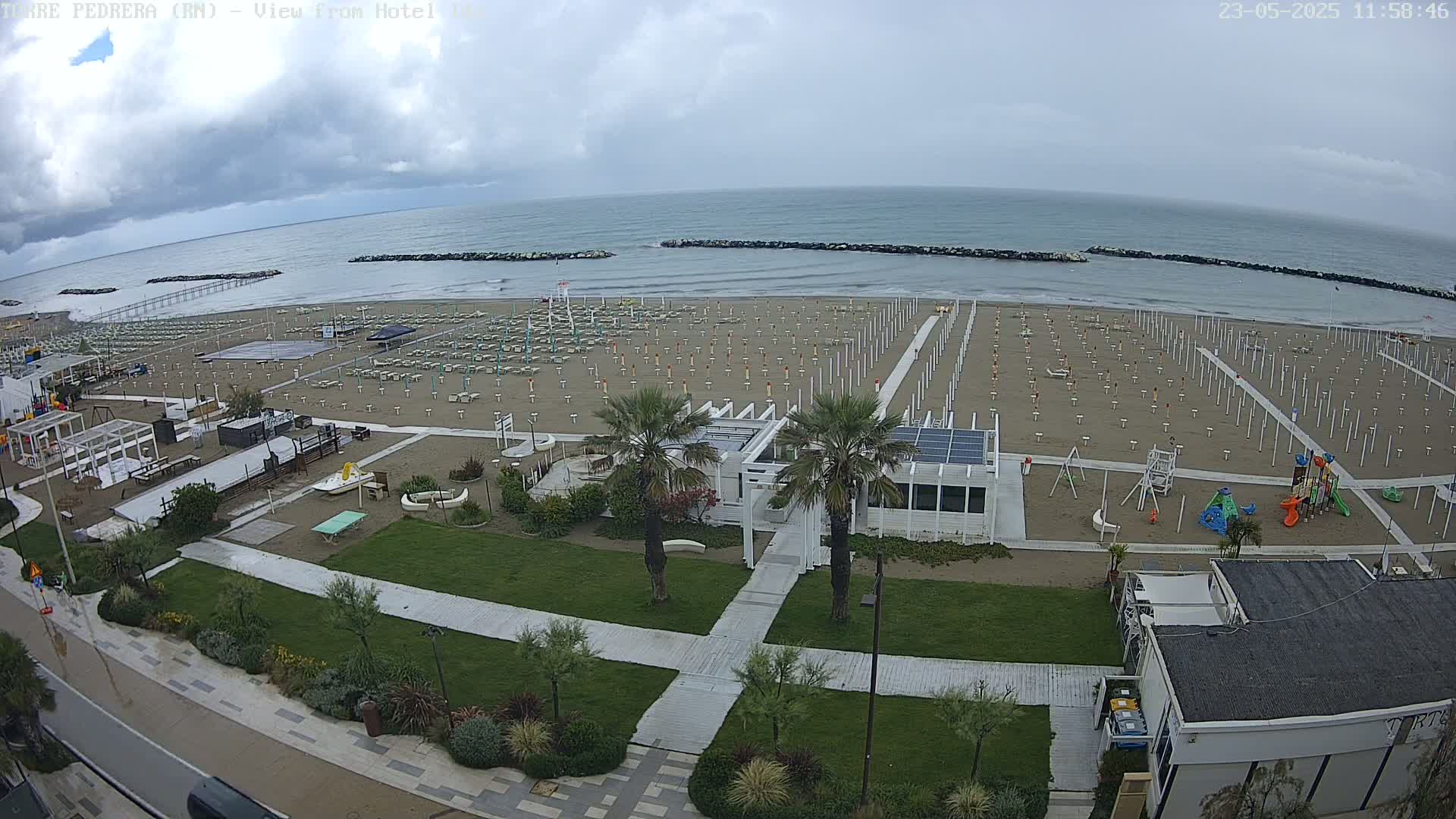 An overhead view of an empty beach with rows of closed umbrellas and beach chairs under an overcast sky, adjacent to a grassy area with landscaping, walkways, and a small playground.