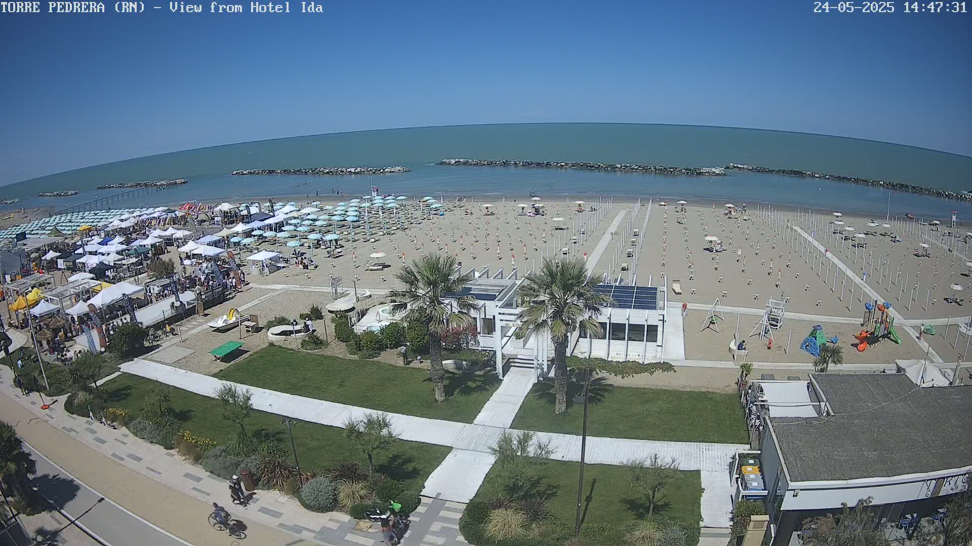 A sandy beach with numerous umbrellas and beachgoers under a clear blue sky is shown, with a calm sea in the background, and a boardwalk leading to a building near a playground.