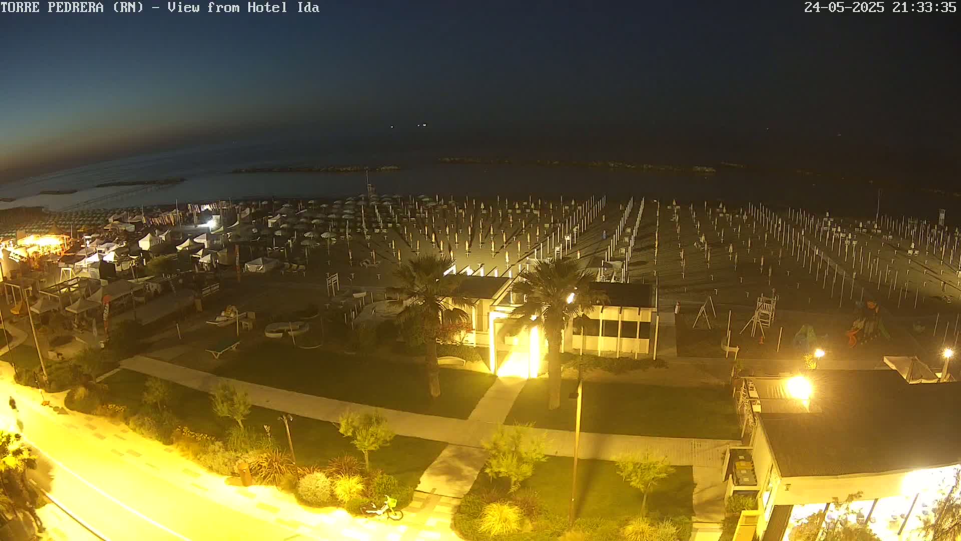 A nighttime view of a beach with rows of closed umbrellas, a building, and a paved walkway, under a dark sky.