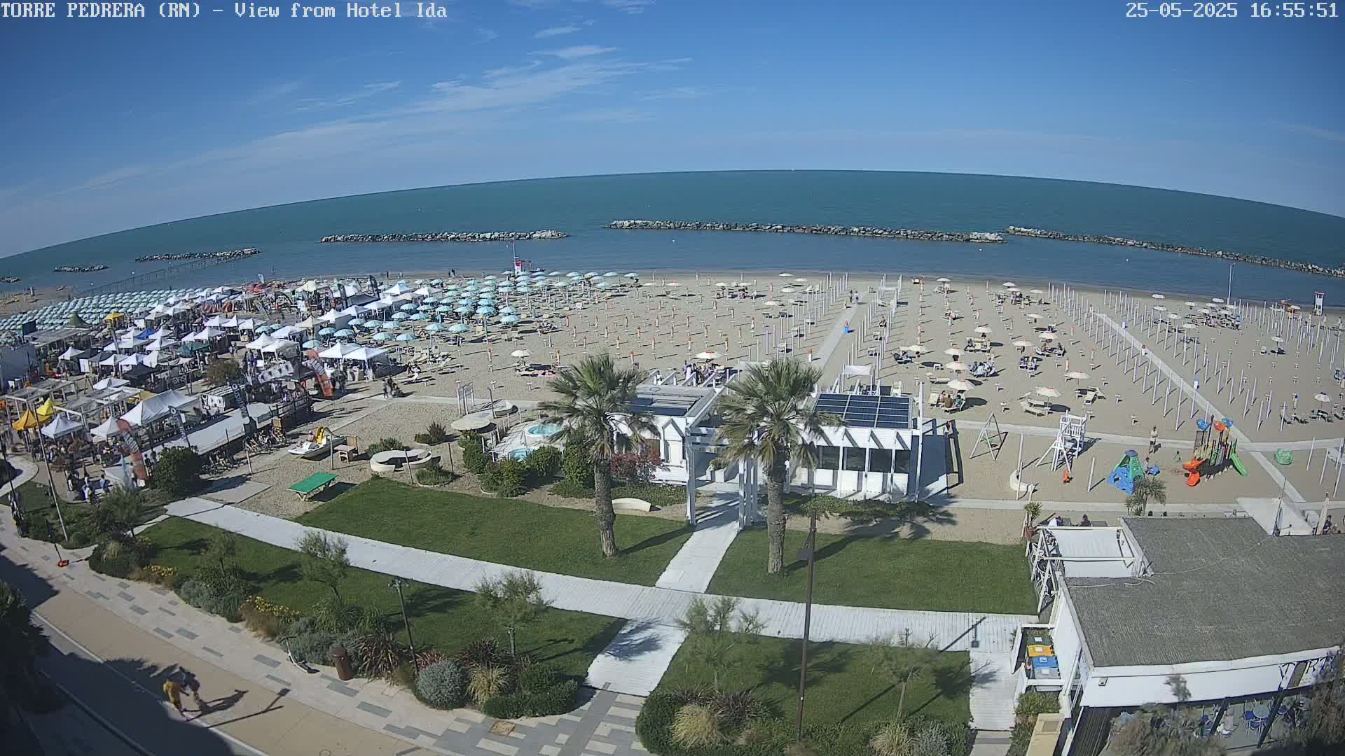 A beach with numerous umbrellas and beachgoers is seen from above on a sunny day, with a calm ocean in the background and a playground near the beachfront buildings.