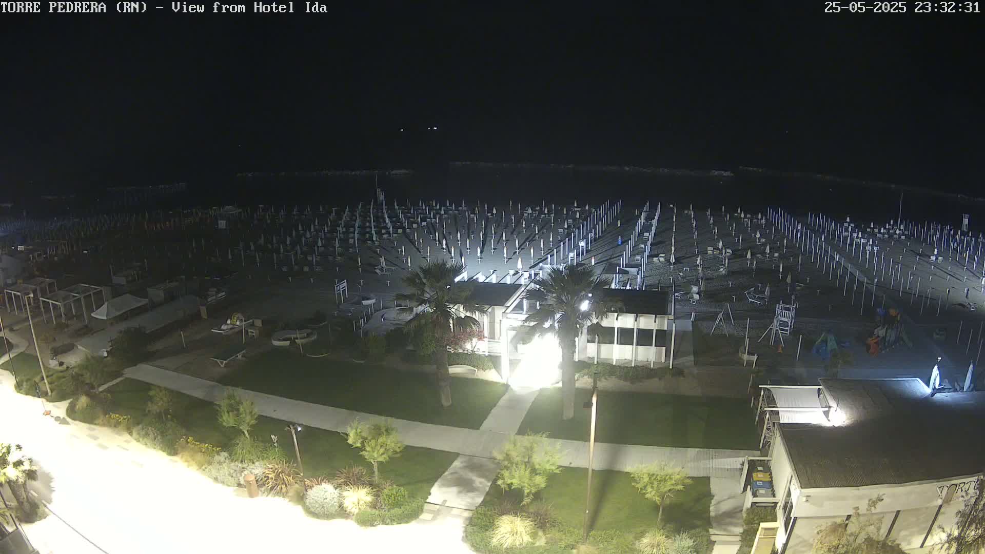 A nighttime, overhead view of a beach with numerous closed umbrellas and beach chairs, a building, and landscaping.