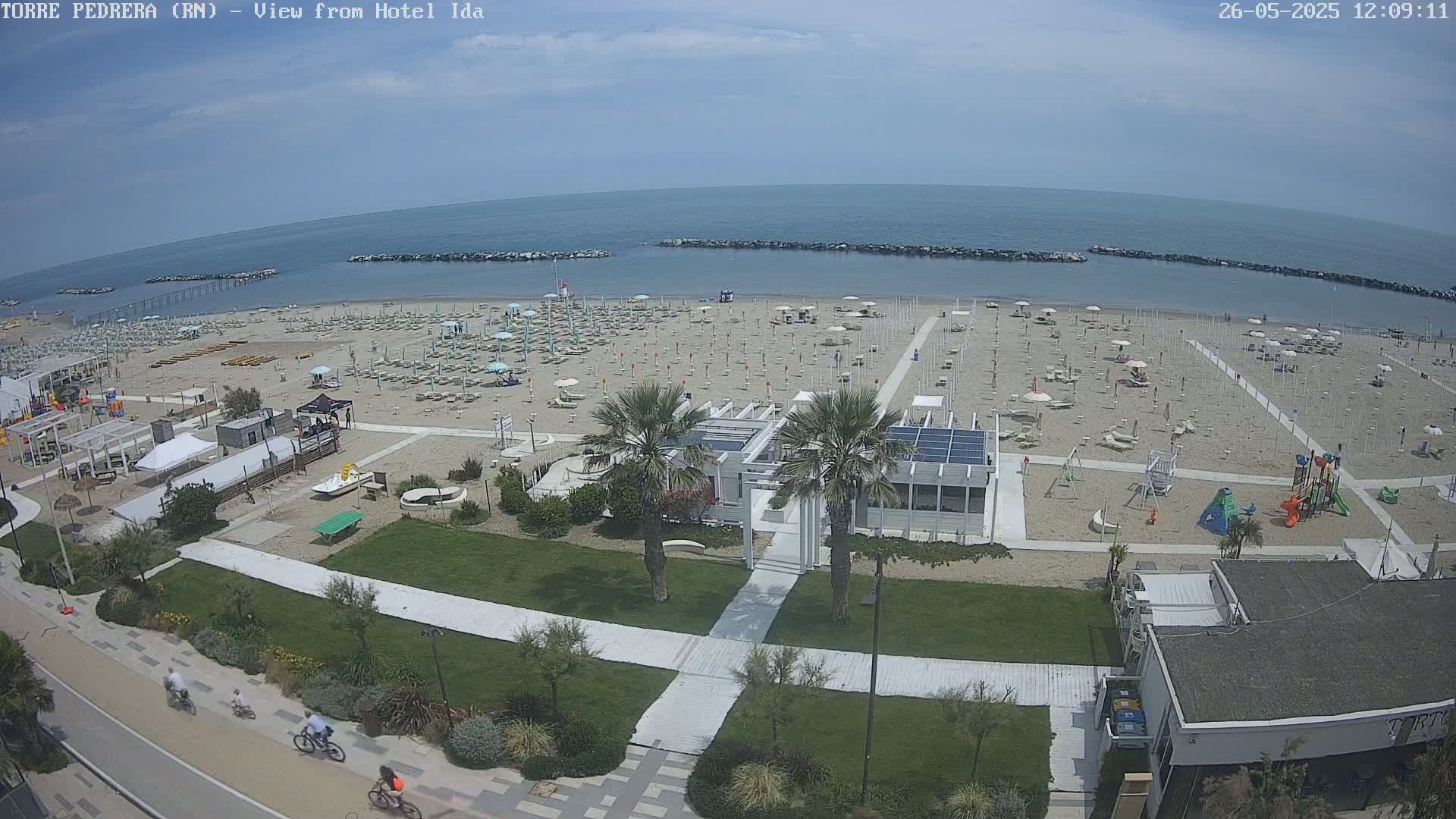 An aerial view shows a beach with numerous umbrellas and lounge chairs, a playground, and a boardwalk, under a mostly sunny sky.