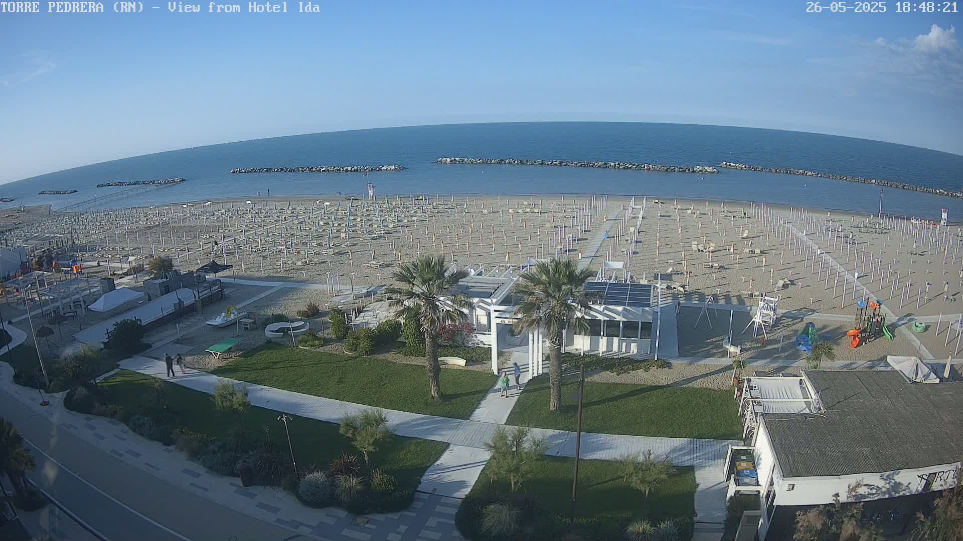 An aerial view of a beach with numerous closed umbrellas and beach chairs under a clear, sunny sky shows a calm sea.