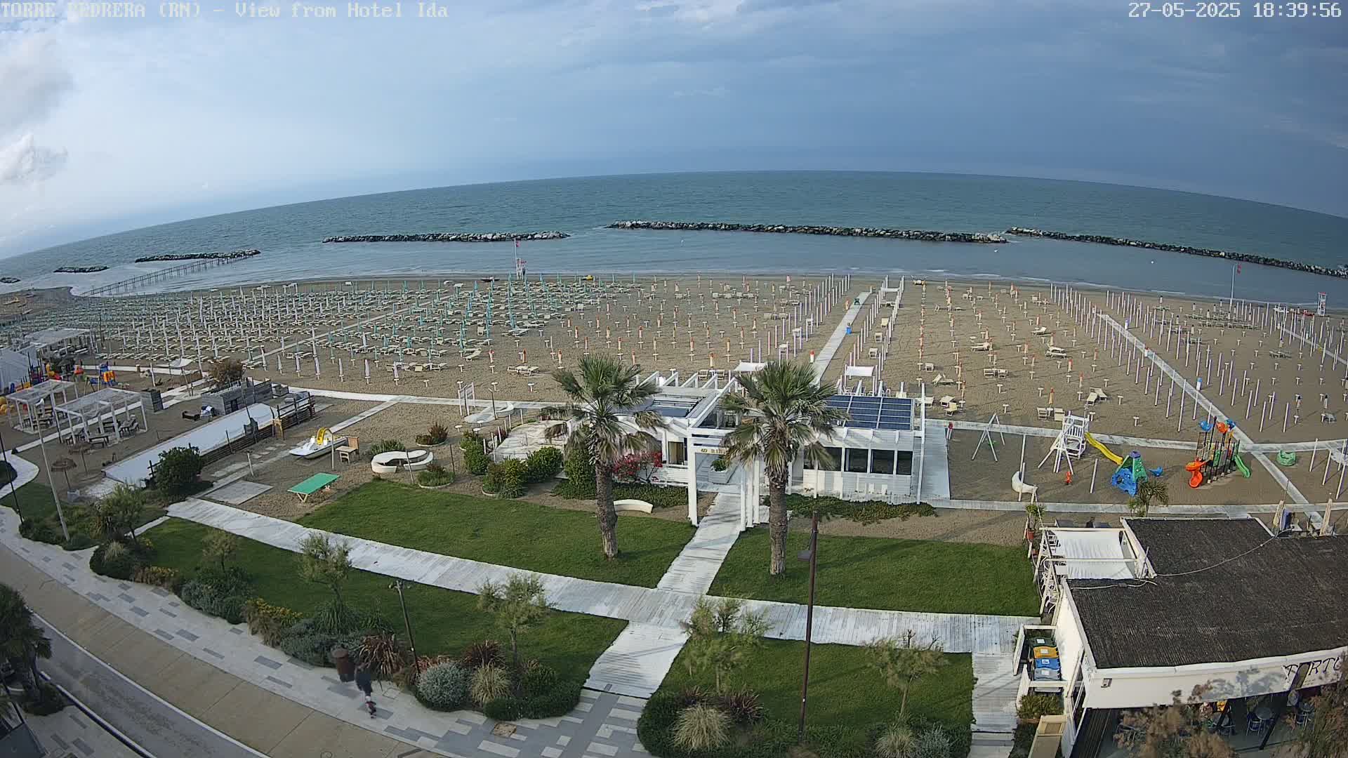 An aerial view shows a mostly empty sandy beach with rows of closed beach umbrellas, a playground, and a beachfront building under a partly cloudy sky.
