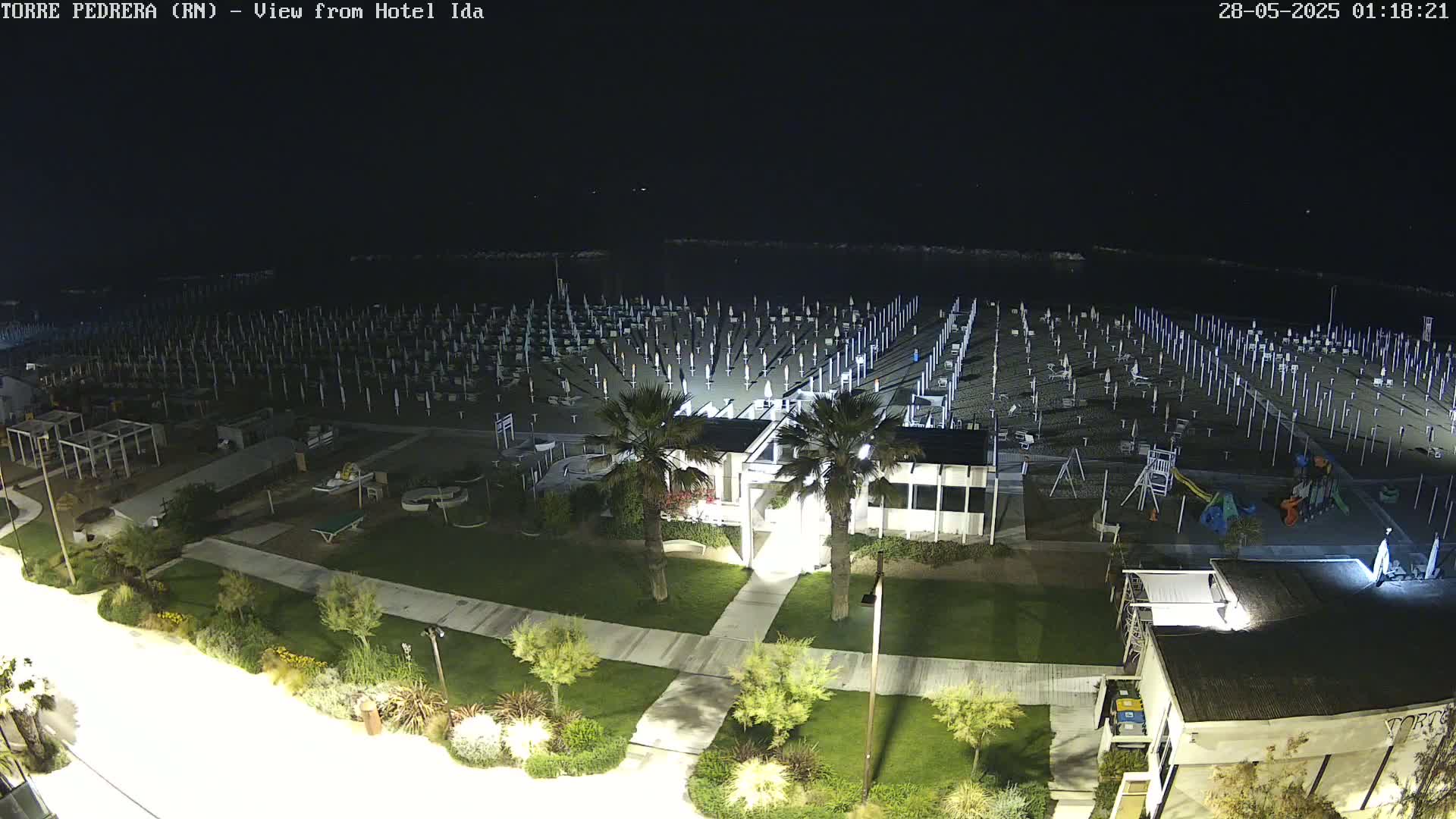 A nighttime, high-angle view of a beach with numerous closed umbrellas and beach chairs, a playground, and some landscaping, all under a dark sky.