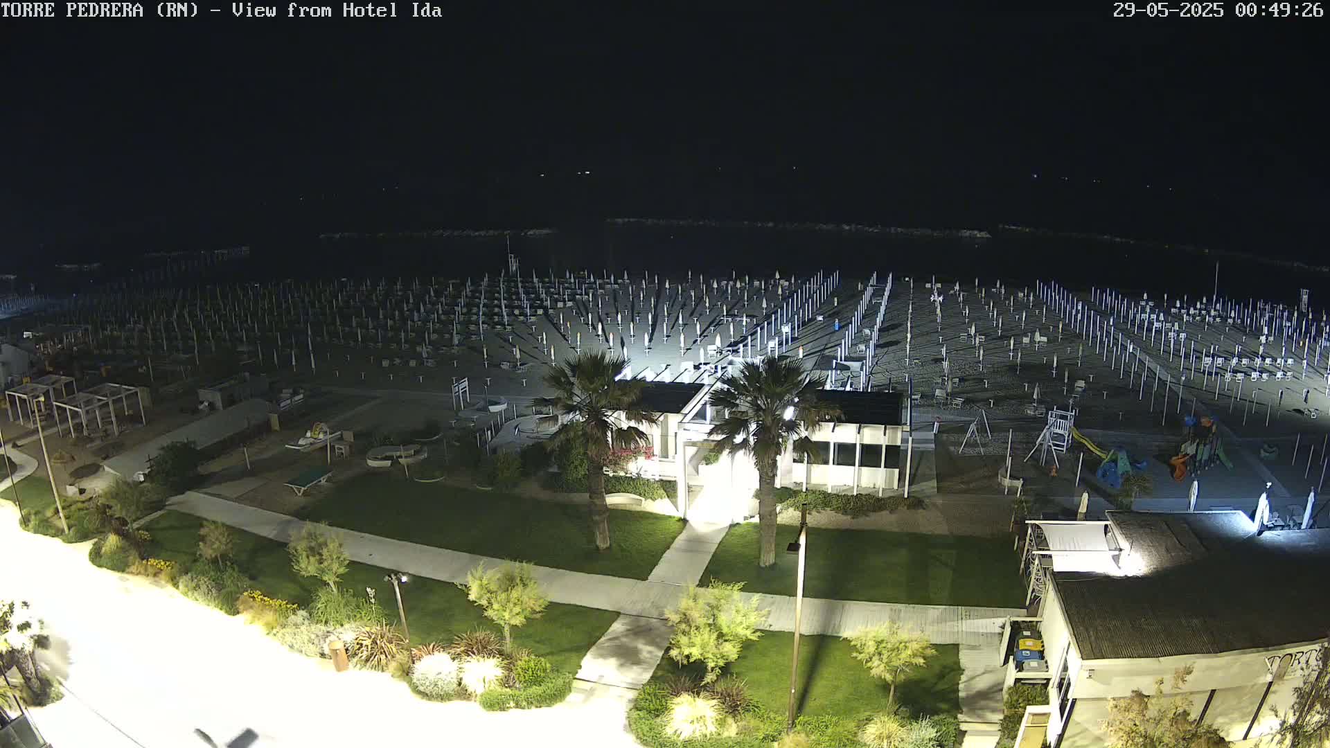 A nighttime view of a beach with rows of closed umbrellas, a playground, and a building, illuminated by artificial lights.