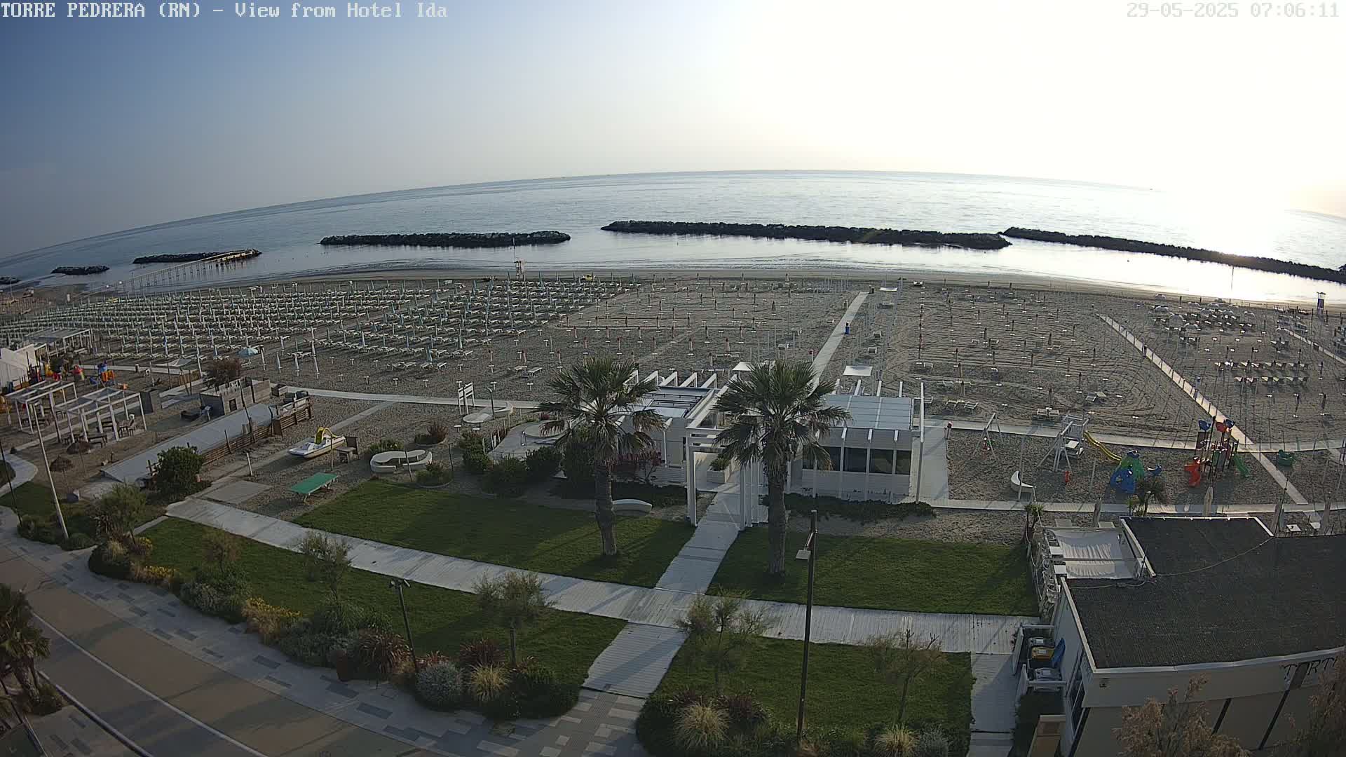 A mostly empty beach with rows of closed umbrellas and beach chairs, a playground, and a beachfront restaurant, all under a clear, bright sky.