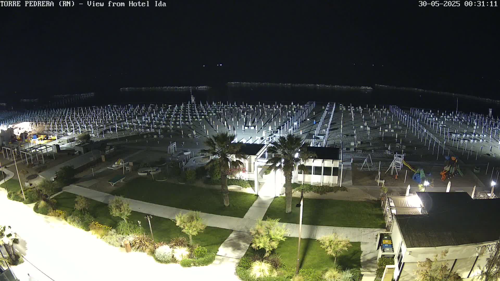 A nighttime, overhead view of a beach with rows of closed umbrellas and beach chairs, a playground, and a building, all illuminated by artificial lights.
