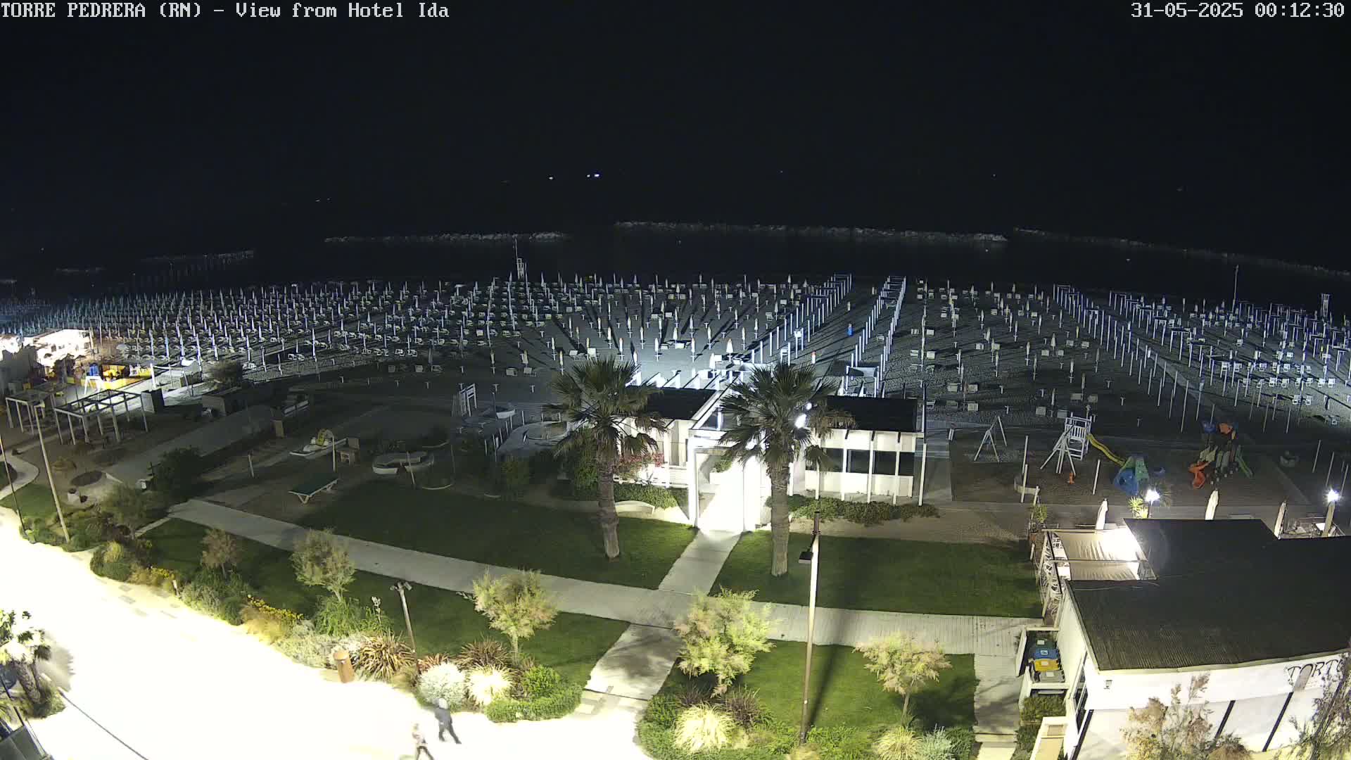A nighttime aerial view of a beach with rows of closed umbrellas and beach chairs, a playground, and some buildings, all illuminated by artificial lights.