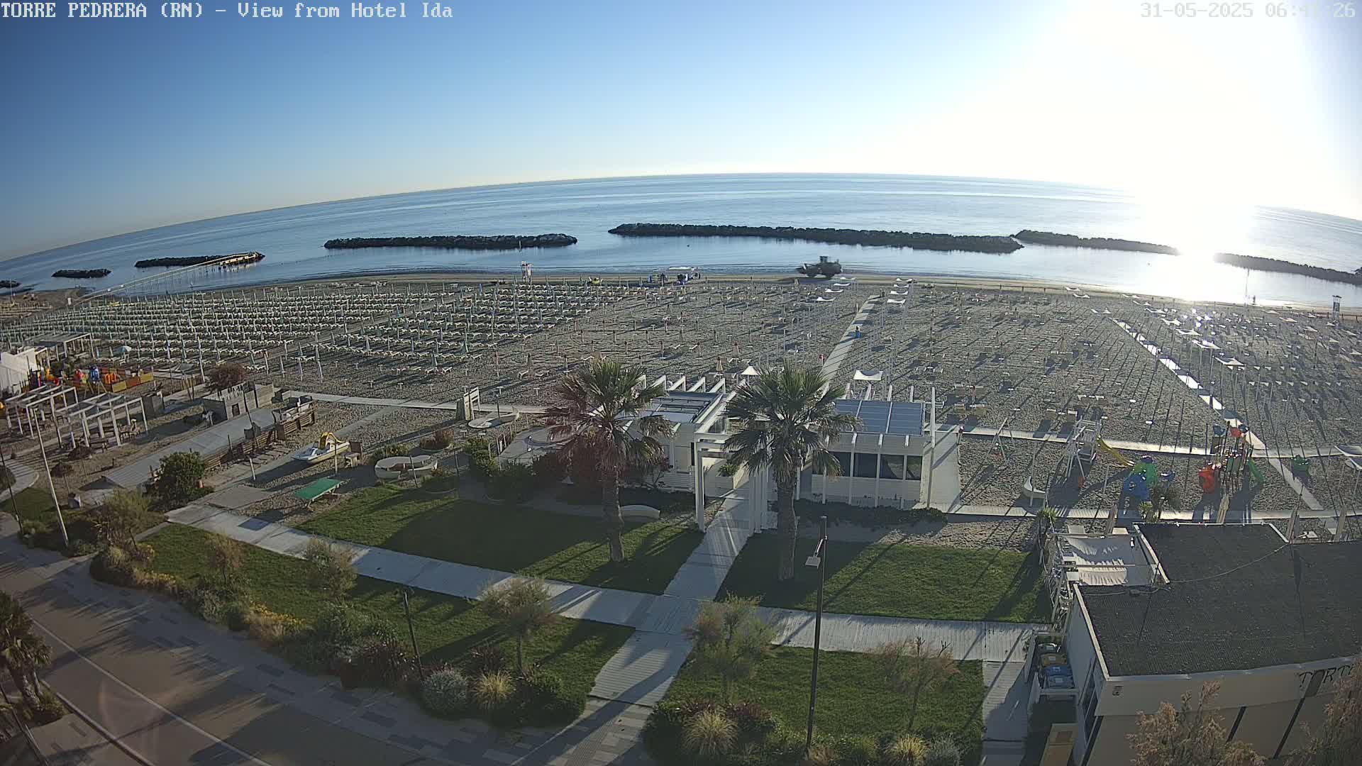 A beach with rows of closed umbrellas and lounge chairs is seen from an elevated view on a sunny morning, with calm water and breakwaters visible in the background.