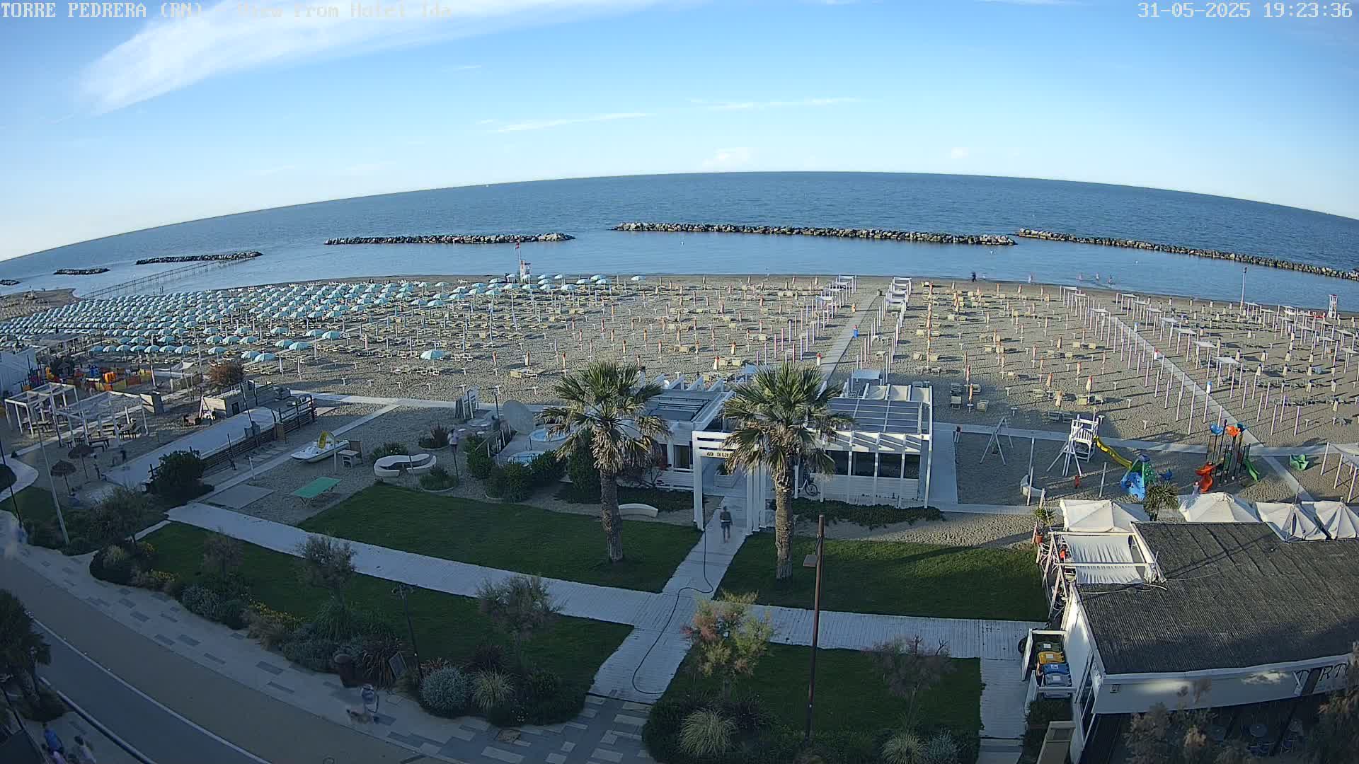 A beach with numerous light-blue umbrellas under a clear blue sky, next to a calm sea with rock breakwaters, and adjacent to a grassy area with palm trees and buildings.