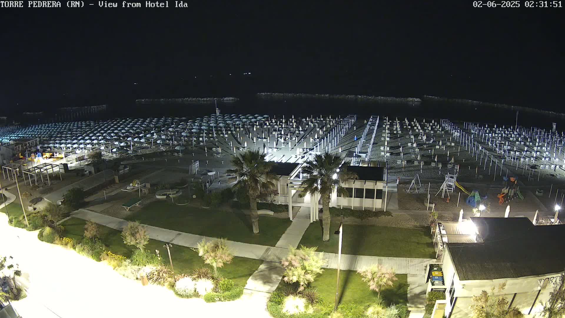 A nighttime, high-angle view of a beach with numerous closed umbrellas, a playground, and surrounding landscaping illuminated by lights.