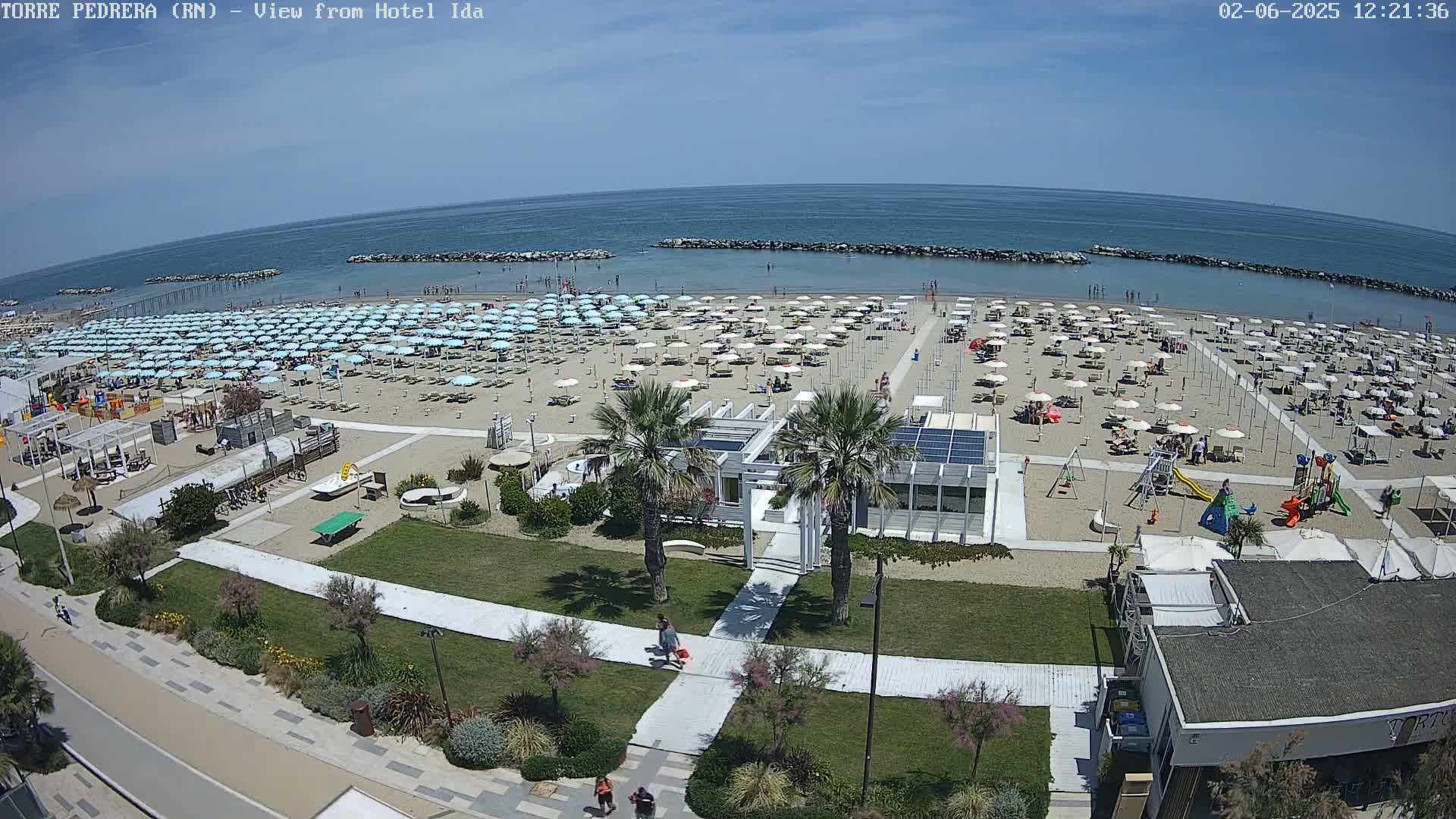 A sunny beach scene shows numerous light-blue beach umbrellas covering a sandy beach next to a calm, blue ocean; a playground and beachside buildings are also visible.