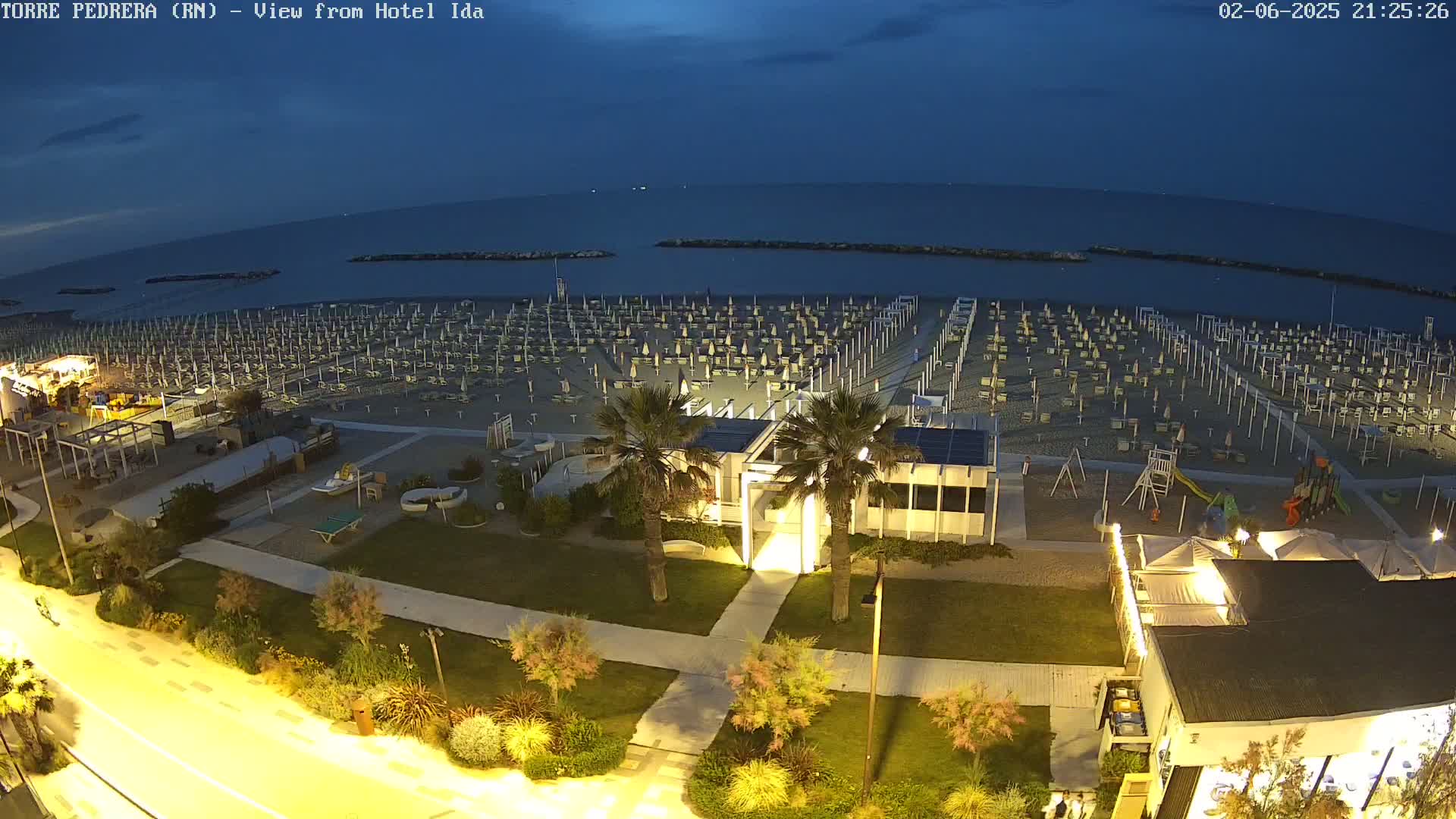 A beach at night, with rows of closed umbrellas and beach chairs, is illuminated, overlooking a calm, dark sea under a twilight sky.