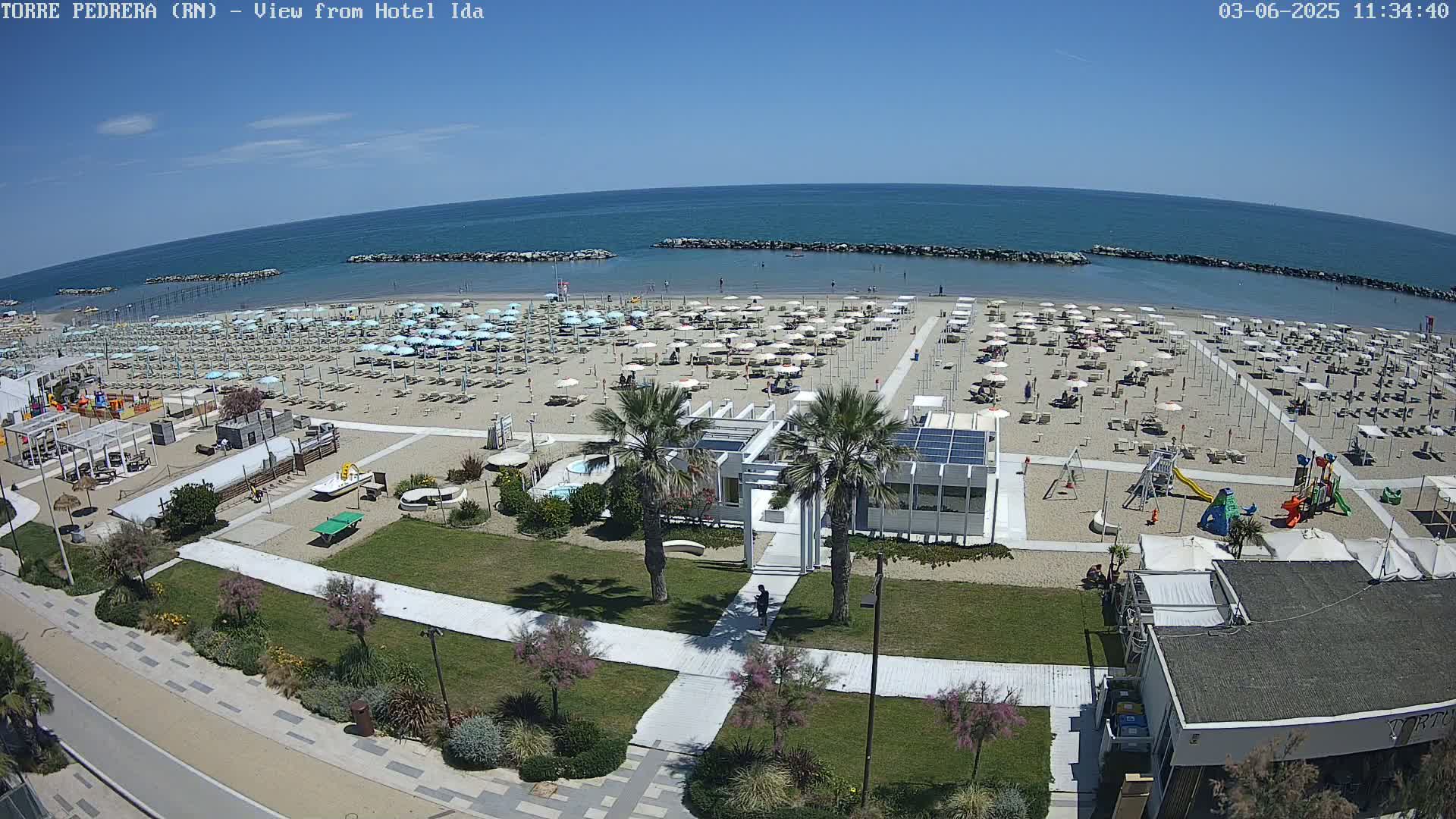 An aerial view shows a sandy beach with numerous umbrellas and beach chairs under a clear blue sky, adjacent to a grassy area with walkways, palm trees, and a playground.