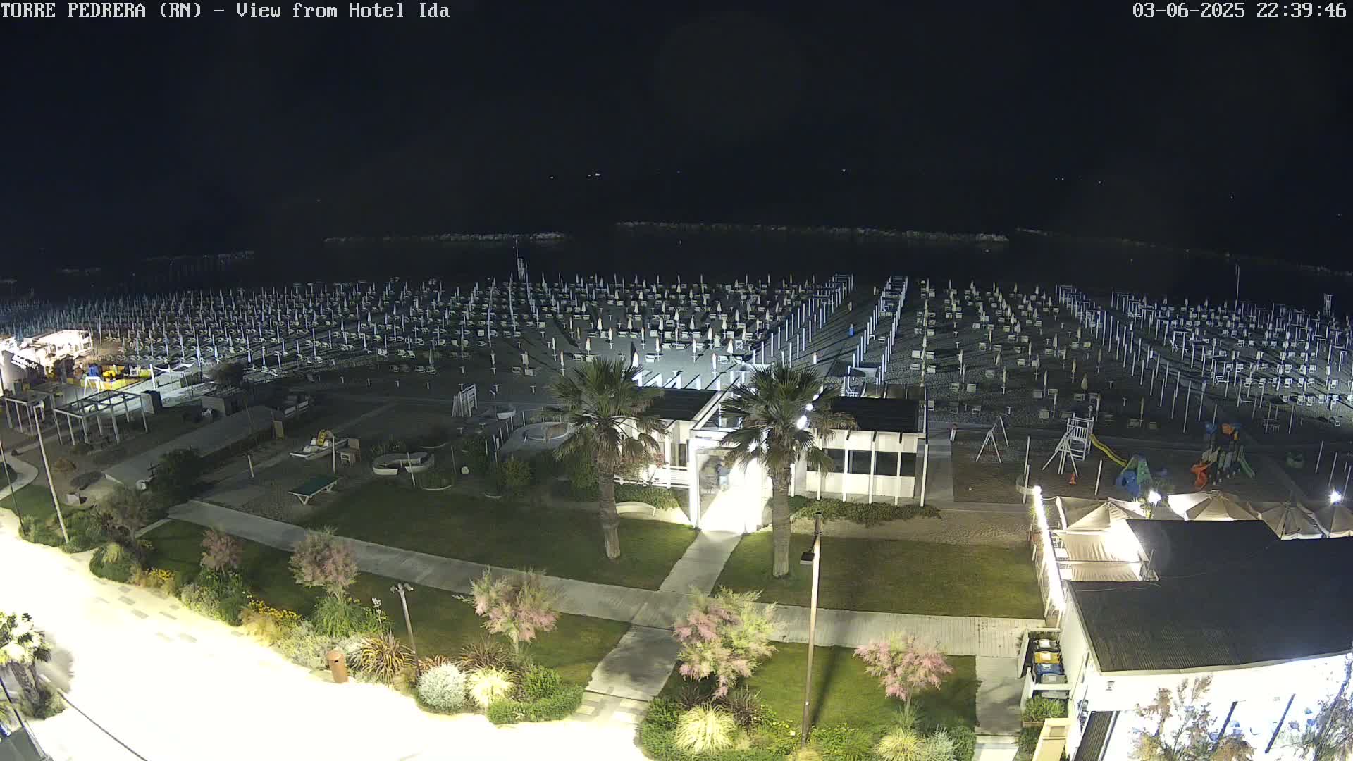 A nighttime view of a beach with rows of closed umbrellas and beach chairs, a playground, and a beachfront building, all illuminated under dark skies.